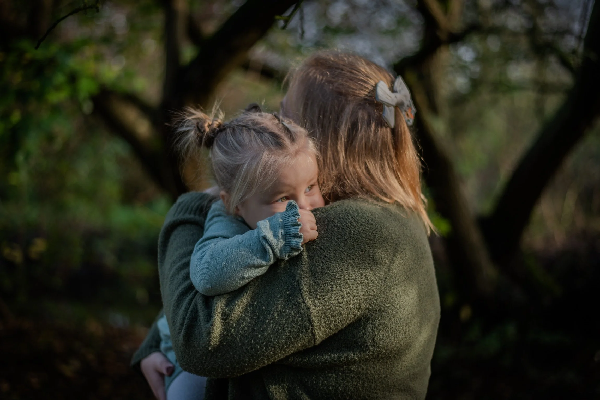 A woman hugging a young girl in an outdoor wooded area. The girl is smiling and looks happy.