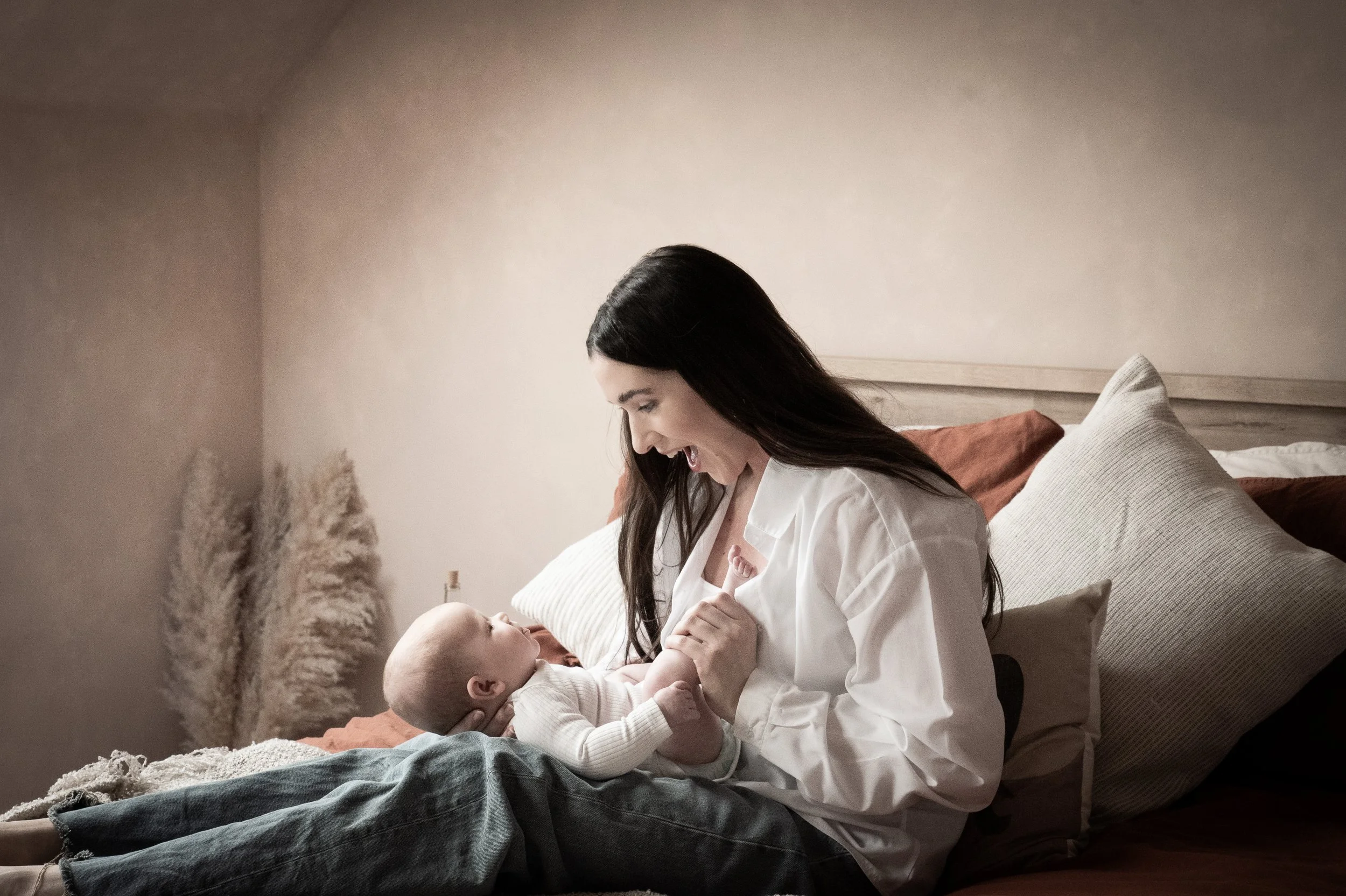 A woman with long dark hair in a white shirt lovingly holding and looking at a baby lying on a bed with pillows.