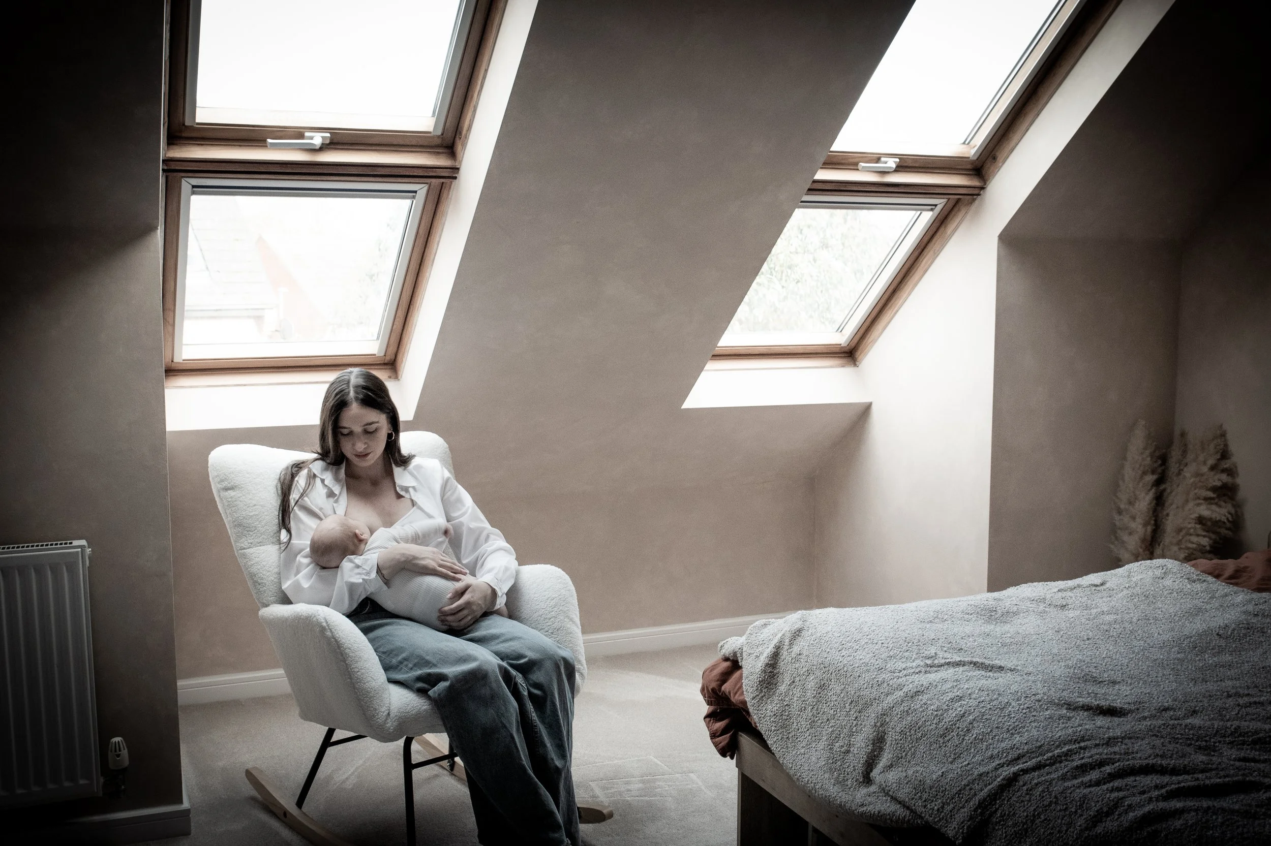A woman sitting in a cream-colored rocking chair breastfeeding a baby in a room with sloped ceilings and three skylight windows.