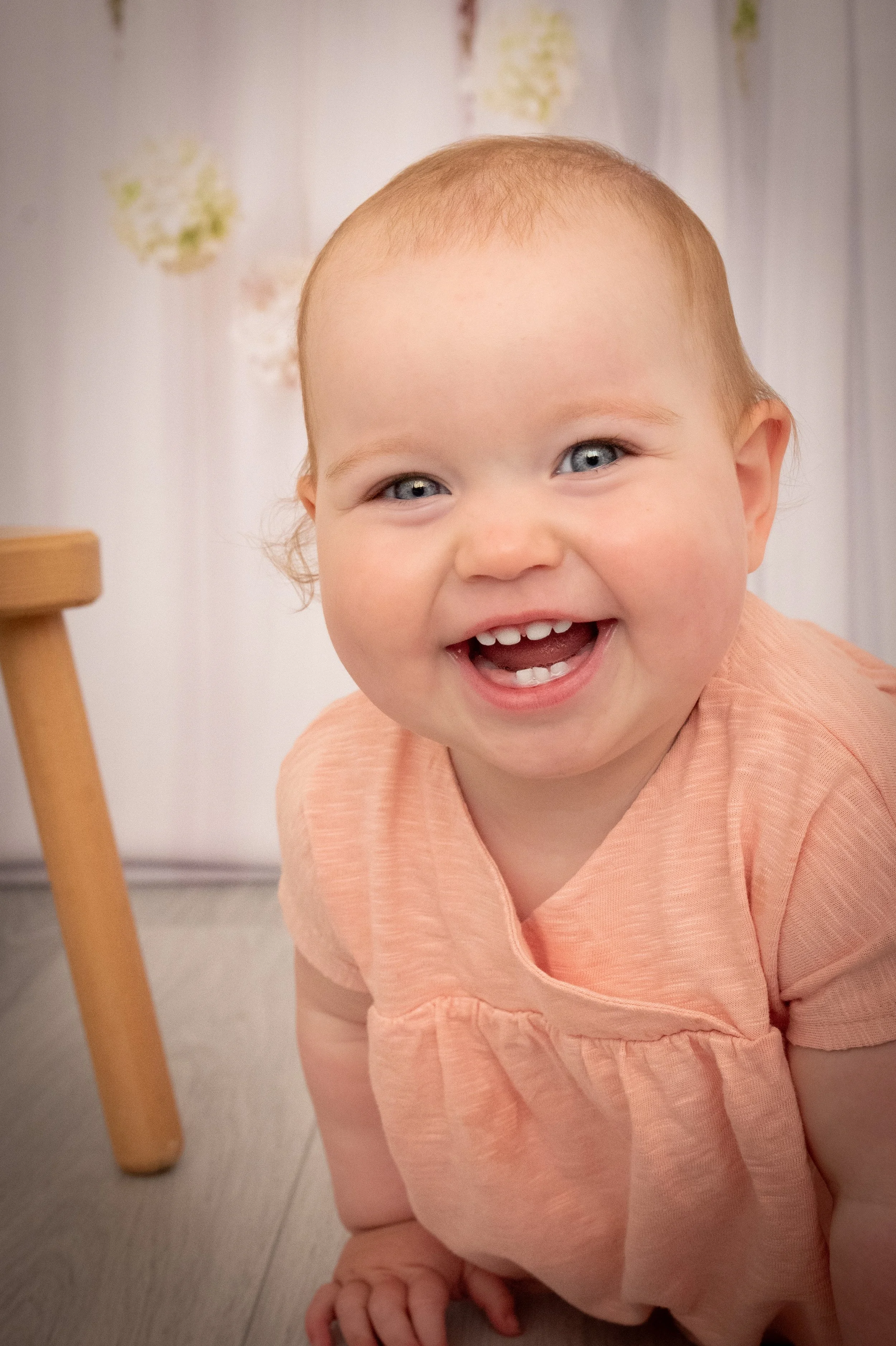 A smiling toddler girl with blue eyes and light brown hair, wearing a peach-colored dress, sitting on the floor with a wooden chair leg visible on the left side of the image.