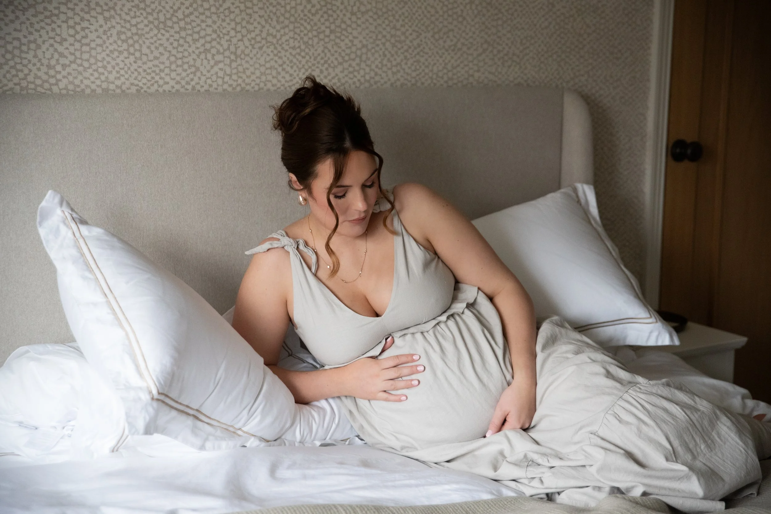 A pregnant woman relaxing on a bed, touching her belly with one hand and looking down, in a bedroom with beige and white decor.
