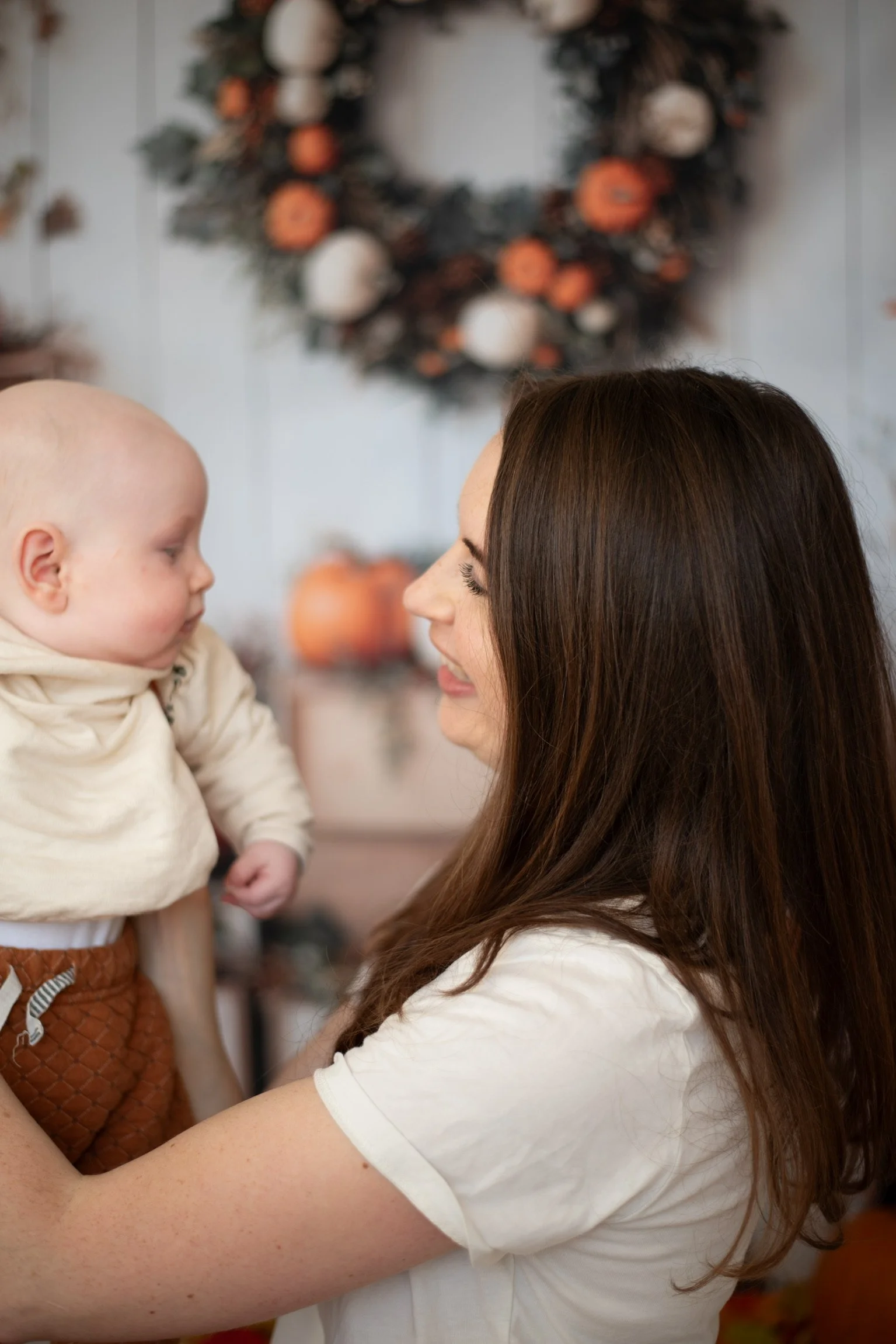 A woman with long brown hair holding a young child with a bald head, against a festive winter background with a decorated wreath and pumpkins.