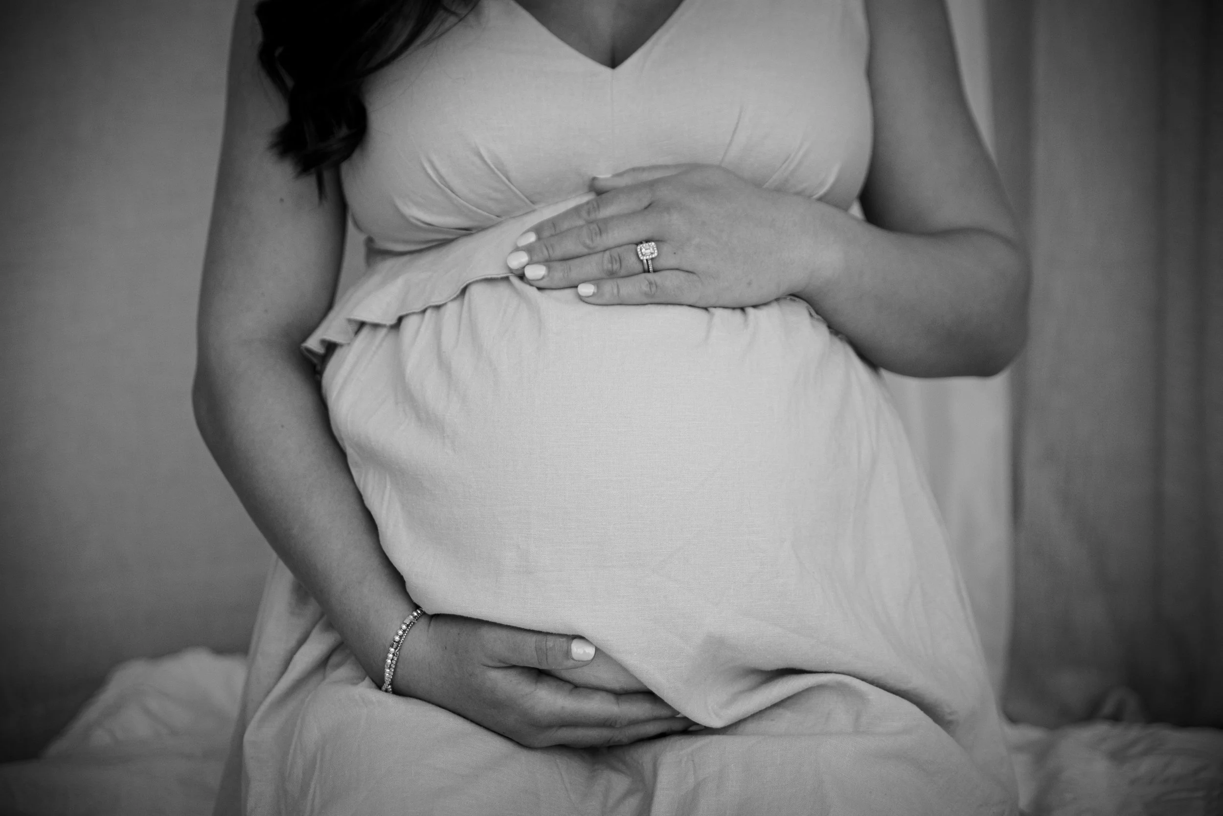 A pregnant woman is sitting on a bed, holding her belly with both hands, wearing a light-colored dress. She has a ring on her finger and a bracelet on her wrist.