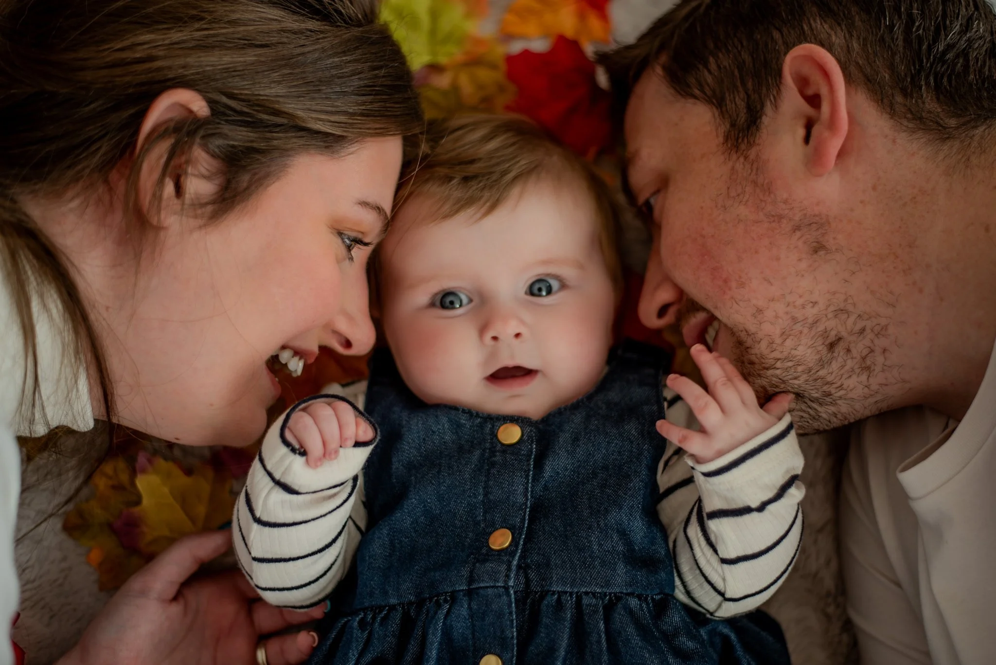 A family of three lying on the floor surrounded by autumn leaves, with the baby in the middle looking surprised, the mother smiling on the left, and the father smiling on the right.