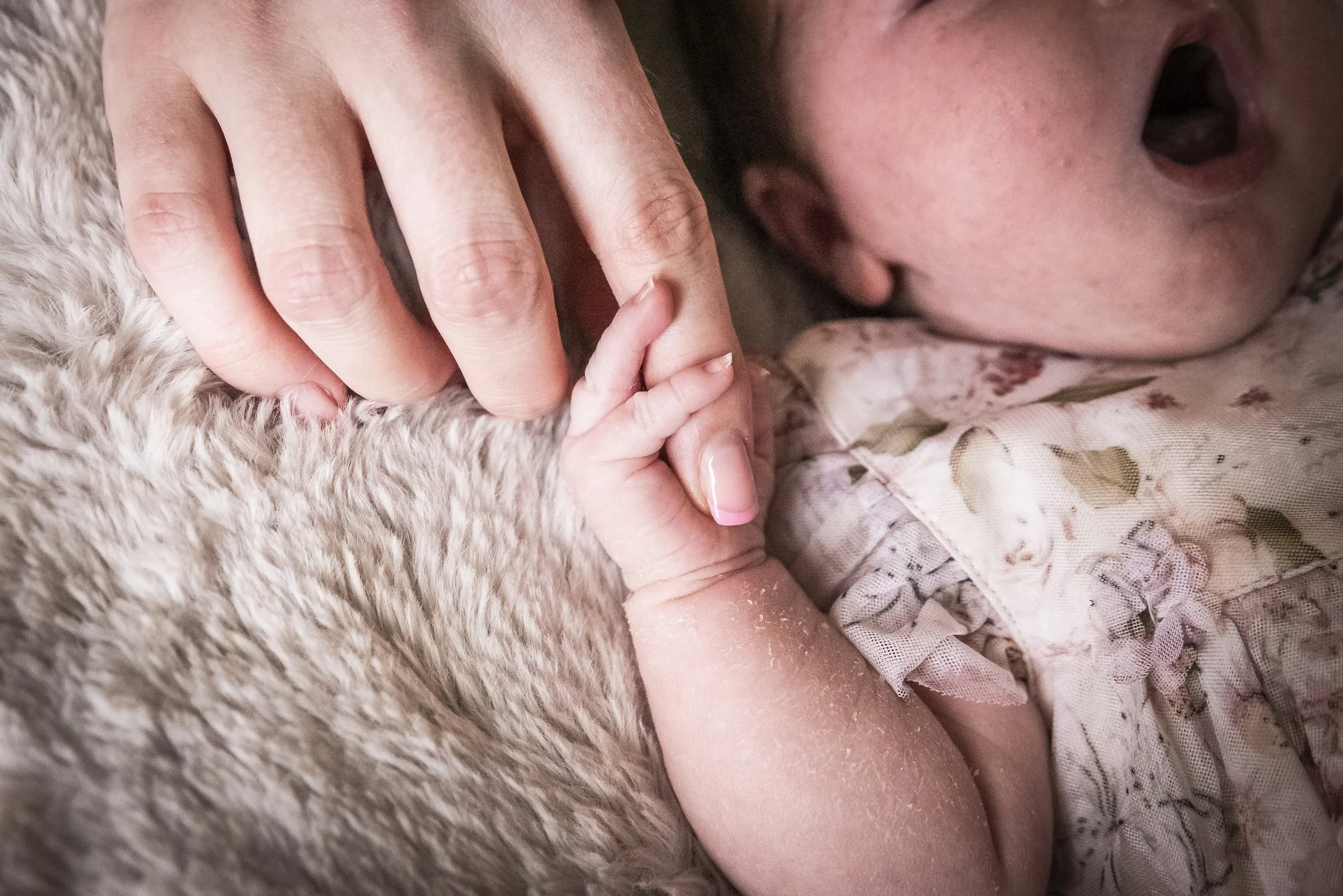 A close-up of an adult's hand gently holding a baby's tiny hand, with the baby's face partially visible and laying on a soft, plush surface.