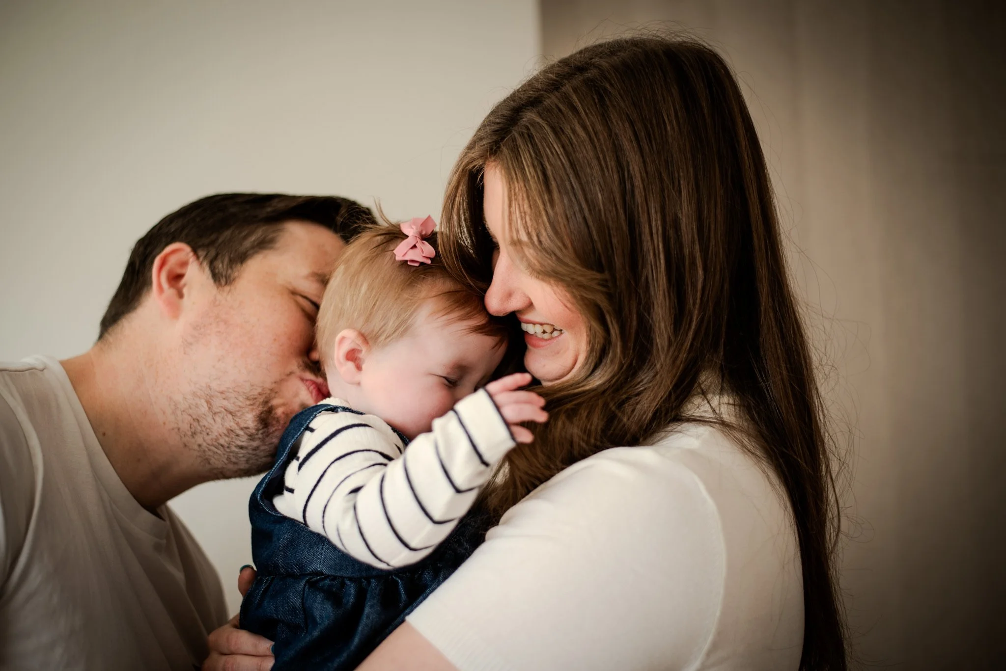 Family of three, parents and toddler, sharing a hug and smiling warmly, indoors.