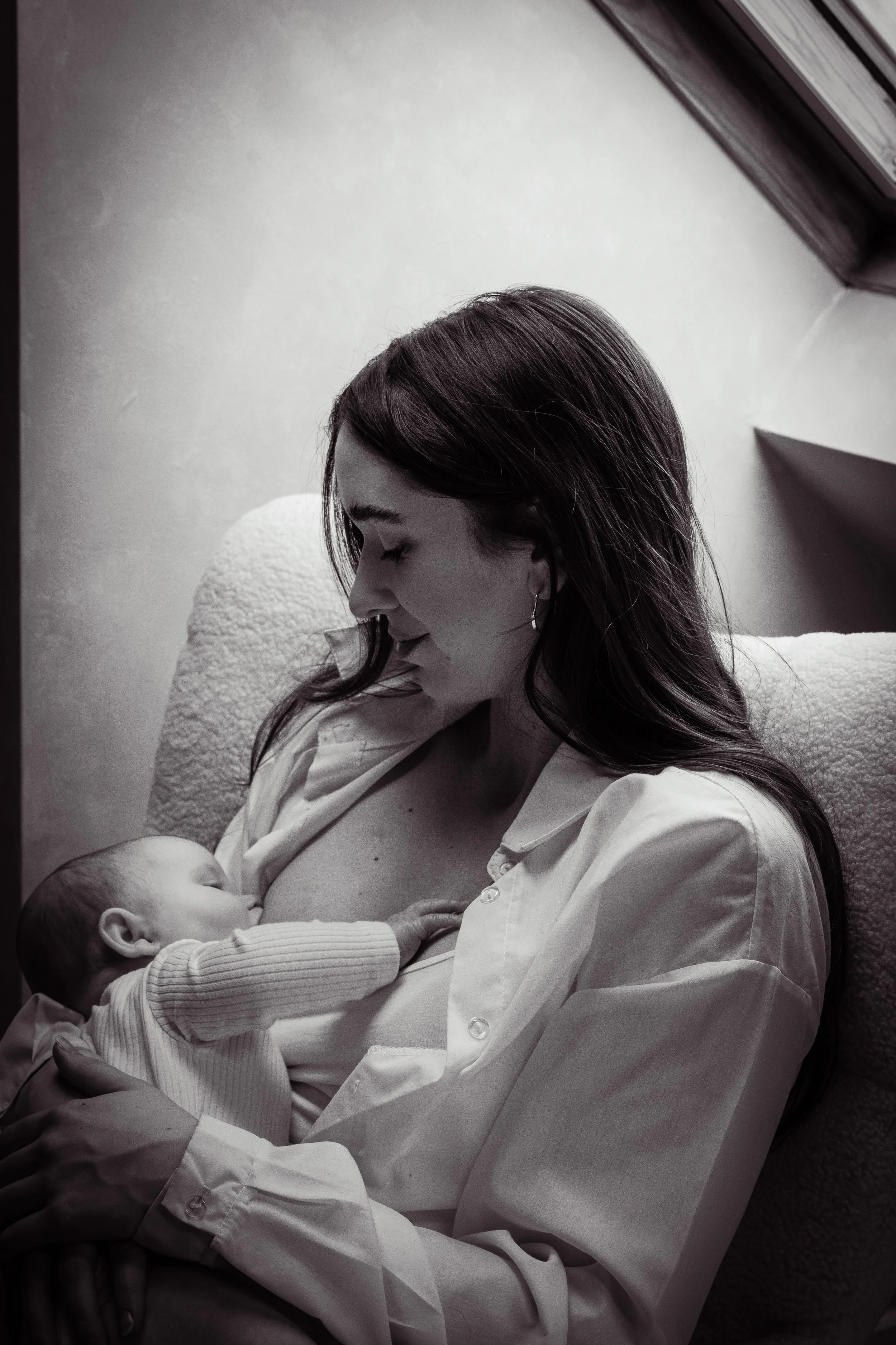 A woman in a white shirt holding a newborn baby on her chest, both sitting on a cozy white chair, with her eyes closed and the baby feeding.