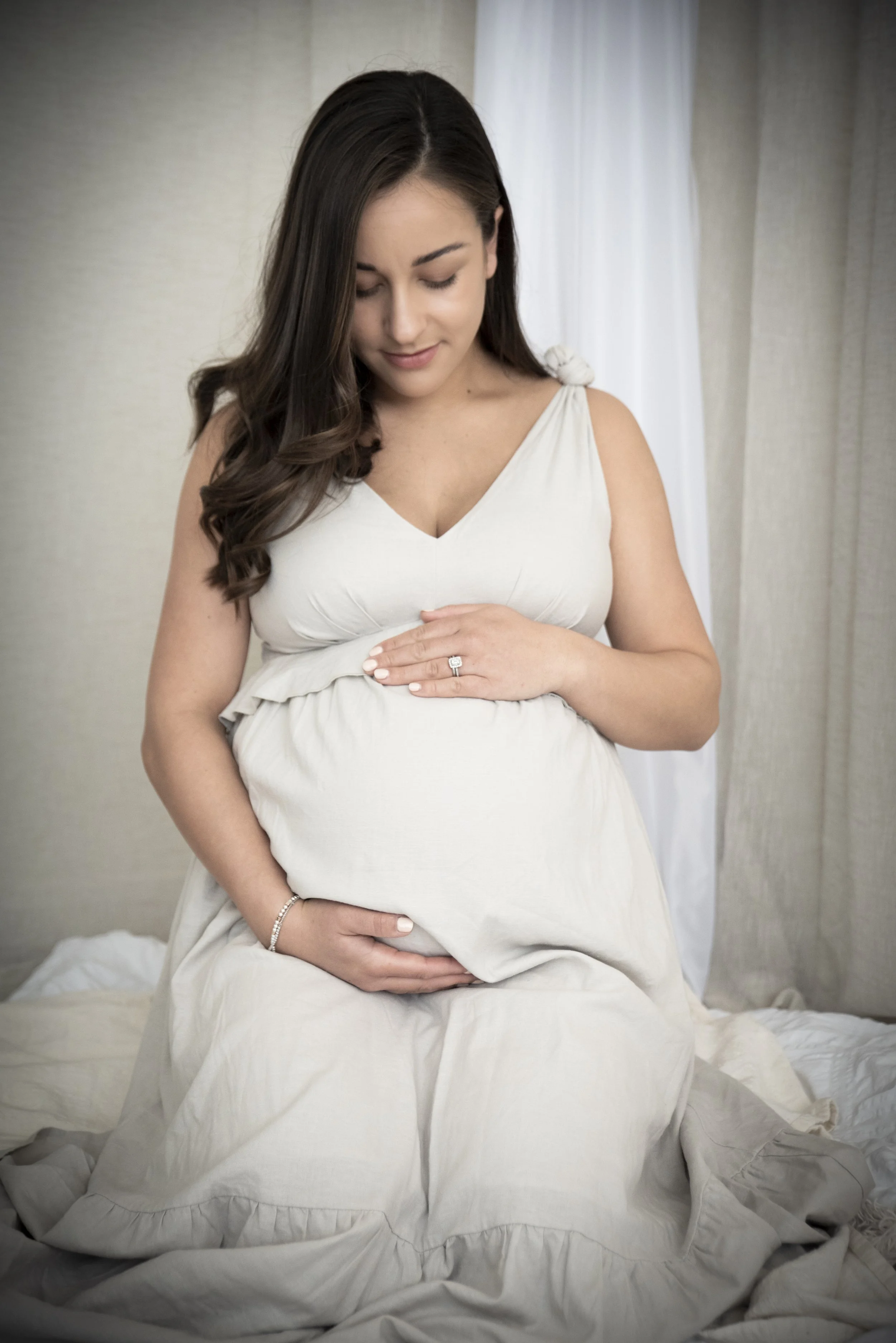 A pregnant woman in a white dress is sitting on a bed, gently holding her belly with both hands and looking down with a serene expression.