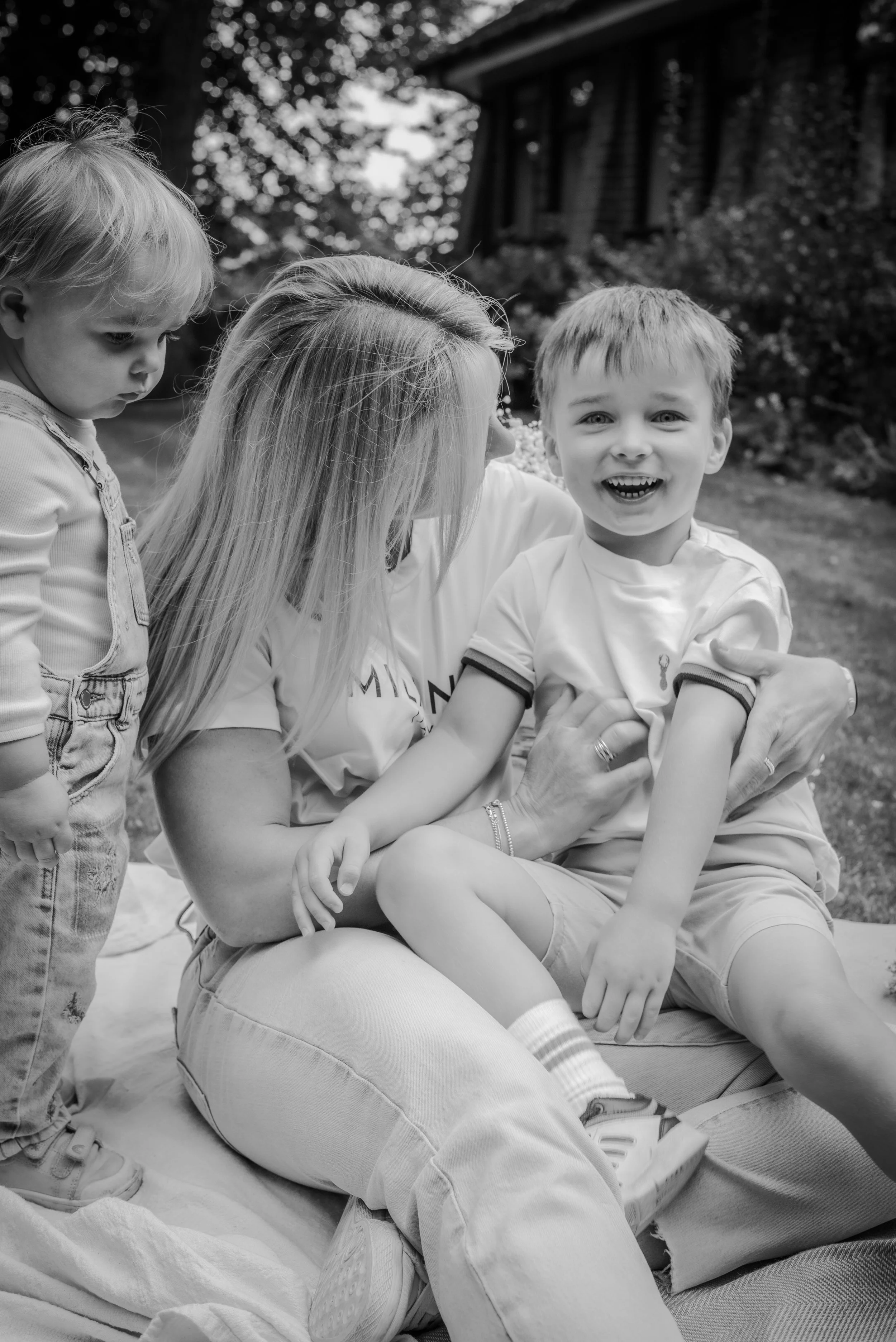 A woman sitting outdoors on a blanket with two young children, one boy smiling at the camera and a girl standing beside her looking down, in a yard with trees and a house in the background.