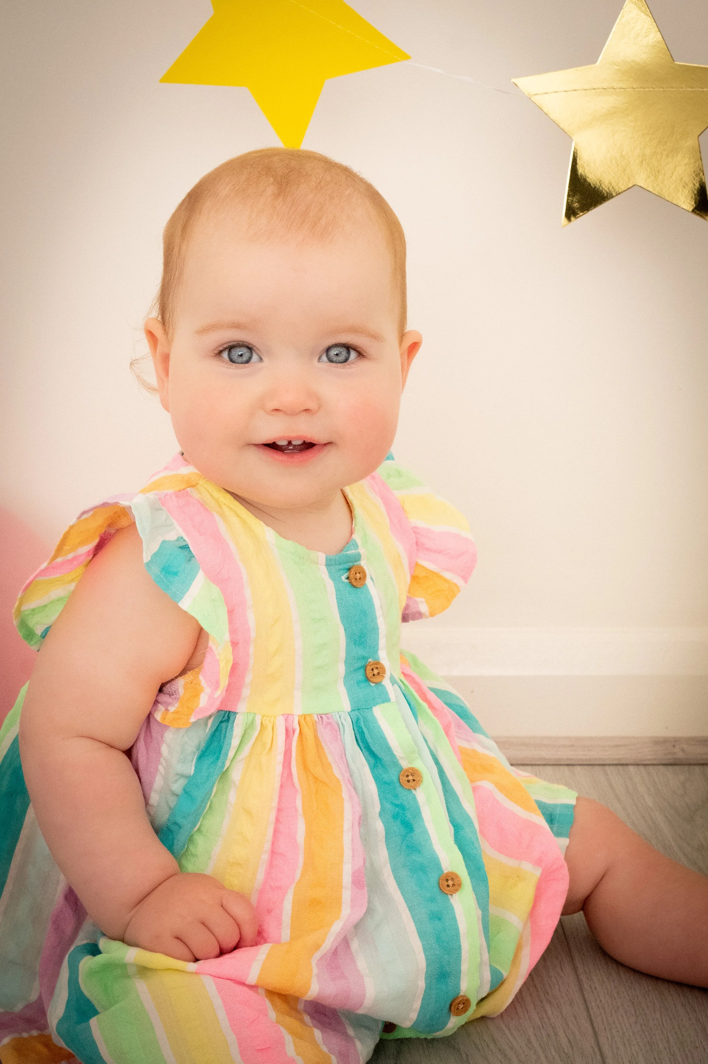 A smiling toddler girl sitting on the floor, wearing a colorful striped dress with brown buttons, and behind her are yellow and gold star decorations.