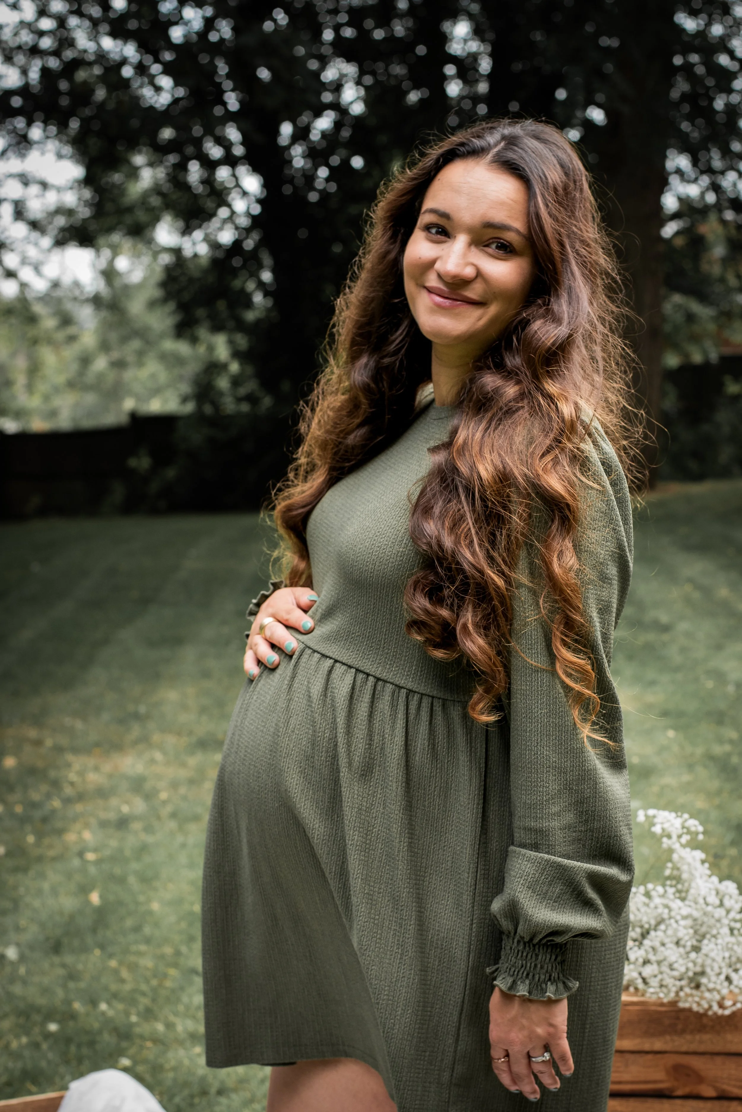 A pregnant woman with long, curly brown hair smiling outdoors in a green dress, holding her belly with her left hand, with trees and grass in the background.