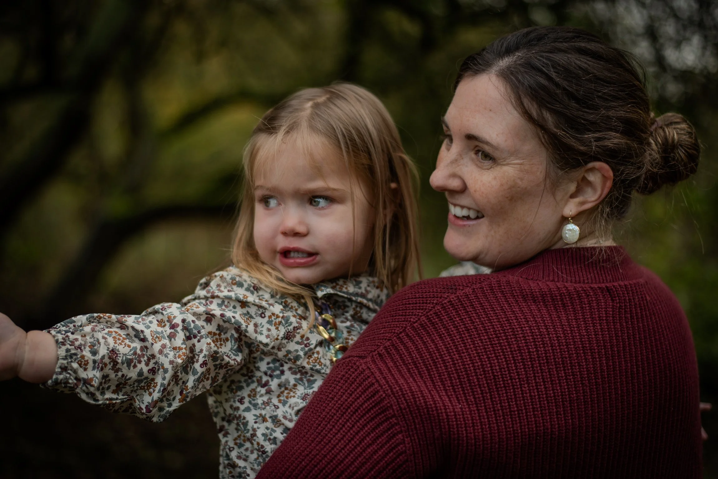 A woman with brown hair in a bun, wearing earrings and a red sweater, smiling while holding a young girl with blonde hair, blue eyes, and a floral jacket, outside in a natural setting.