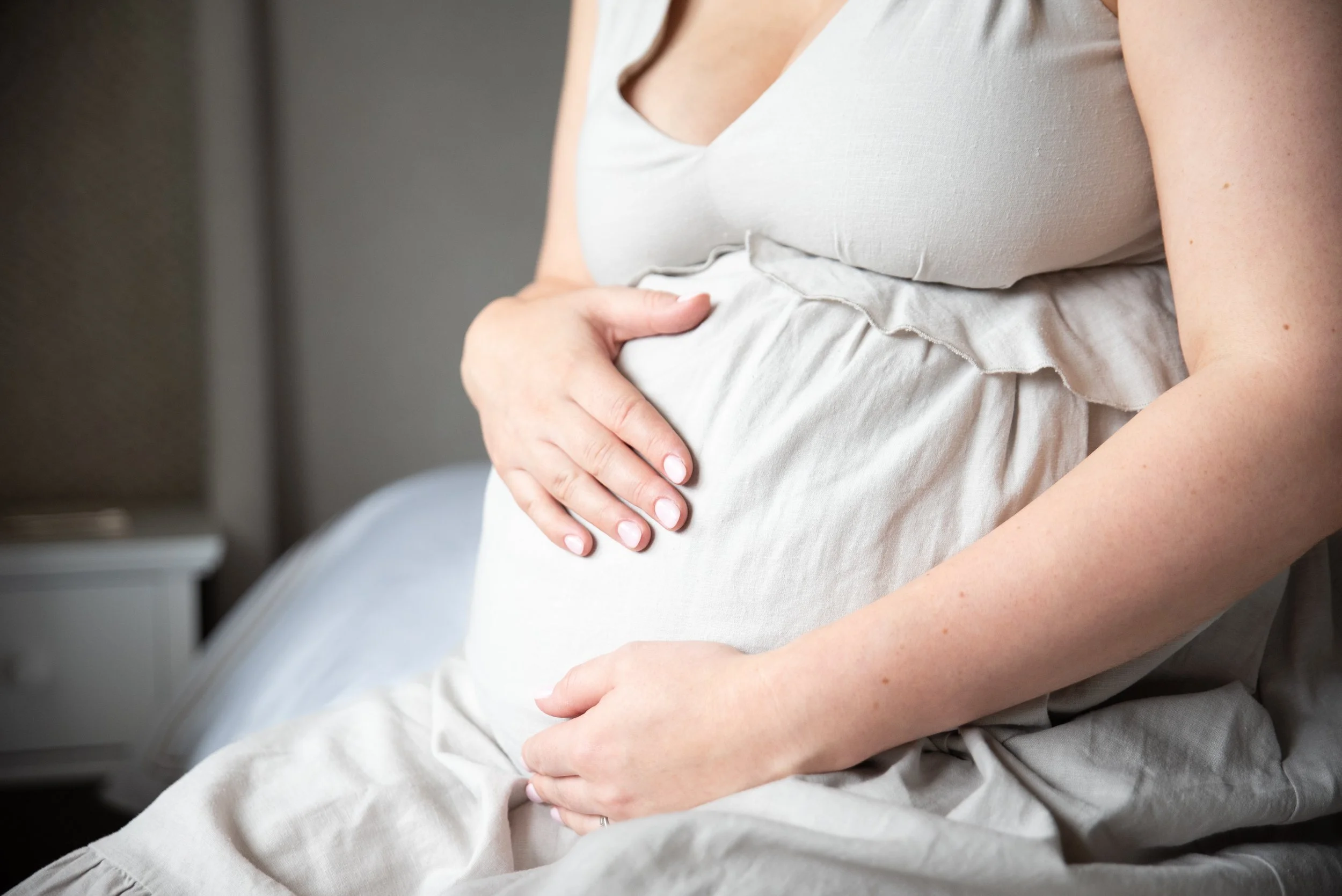 Pregnant woman resting her hand on her belly, sitting on a bed in a bedroom