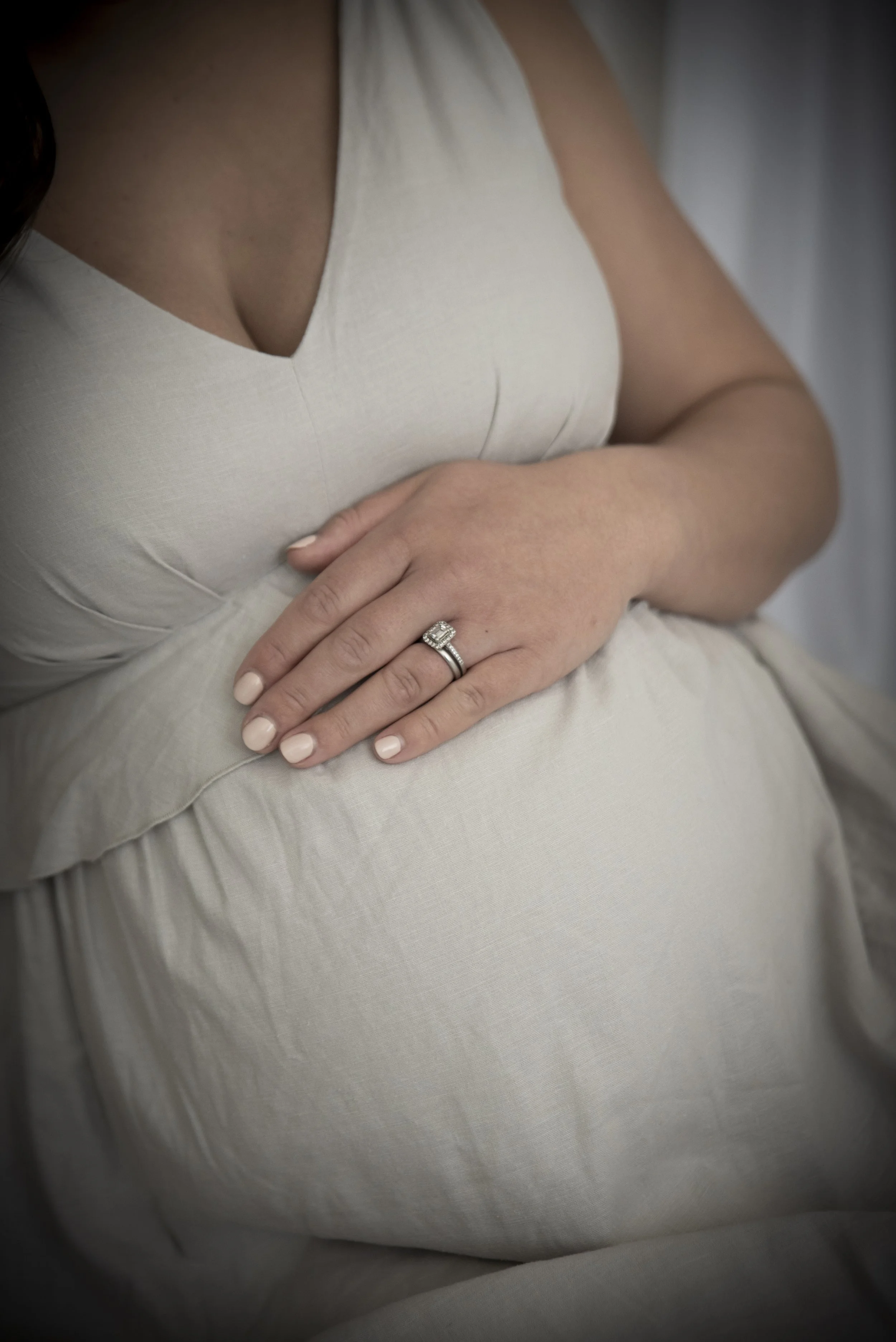 A pregnant woman wearing a cream-colored dress, showing her hand with an engagement ring resting on her belly.