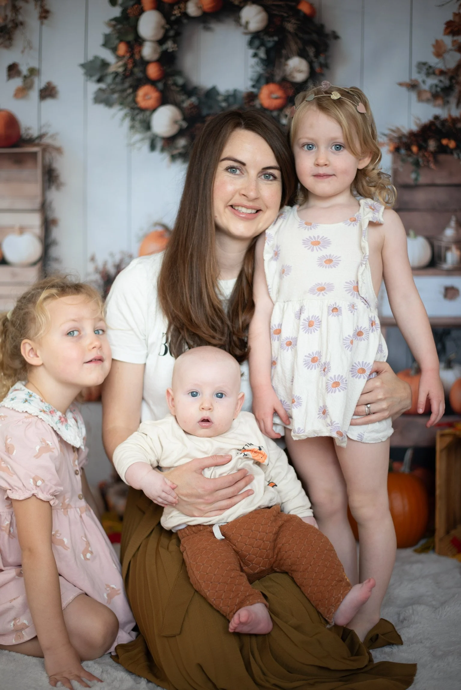 A woman with three young children in front of fall-themed decorations, including pumpkins and a circular fall wreath on a wall.