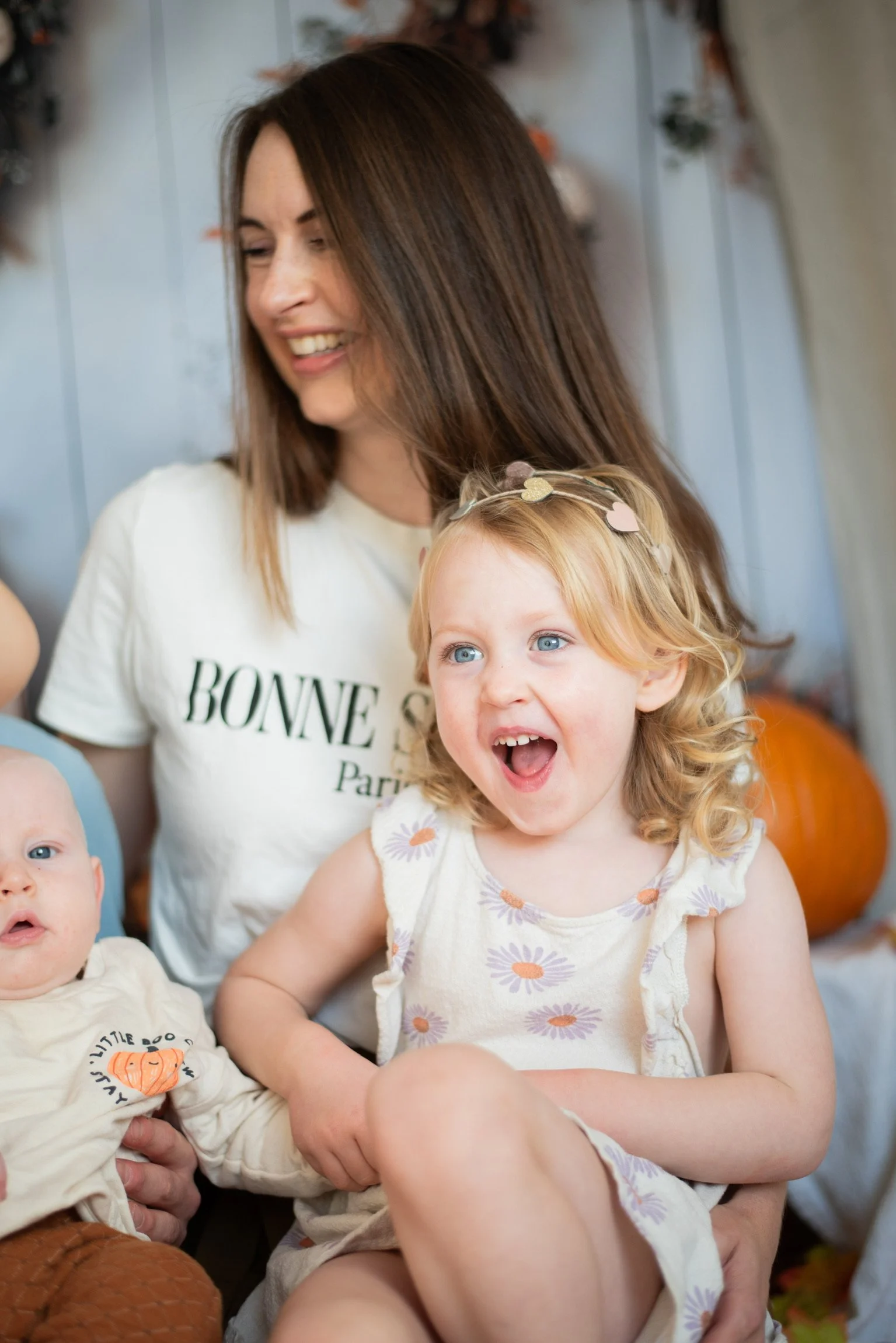 A woman with brown hair smiling, sitting with two young children, a girl with curly blonde hair and blue eyes, and a baby with light hair, in a festive setting with pumpkins and fall decorations.