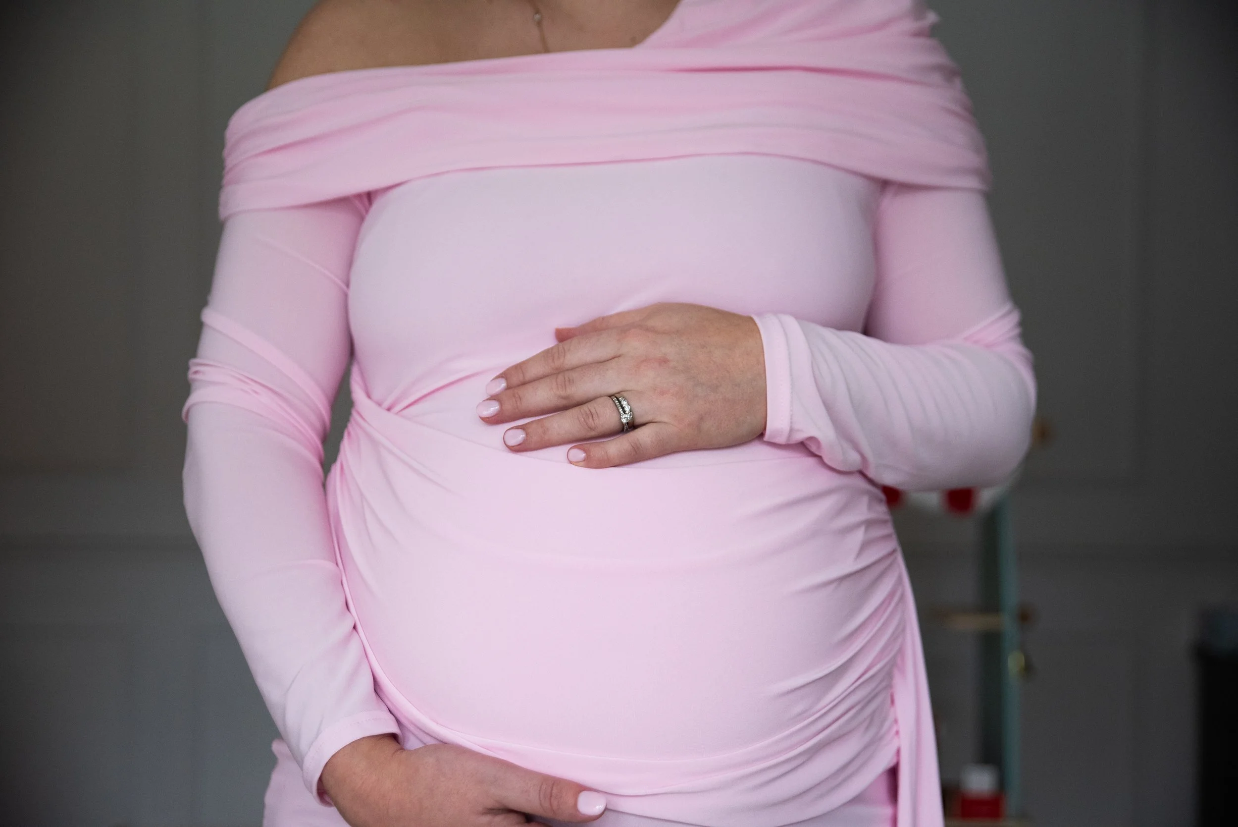 Close-up of a pregnant woman's belly with her hand resting on it, wearing a pink dress with long sleeves and a ring on her finger.