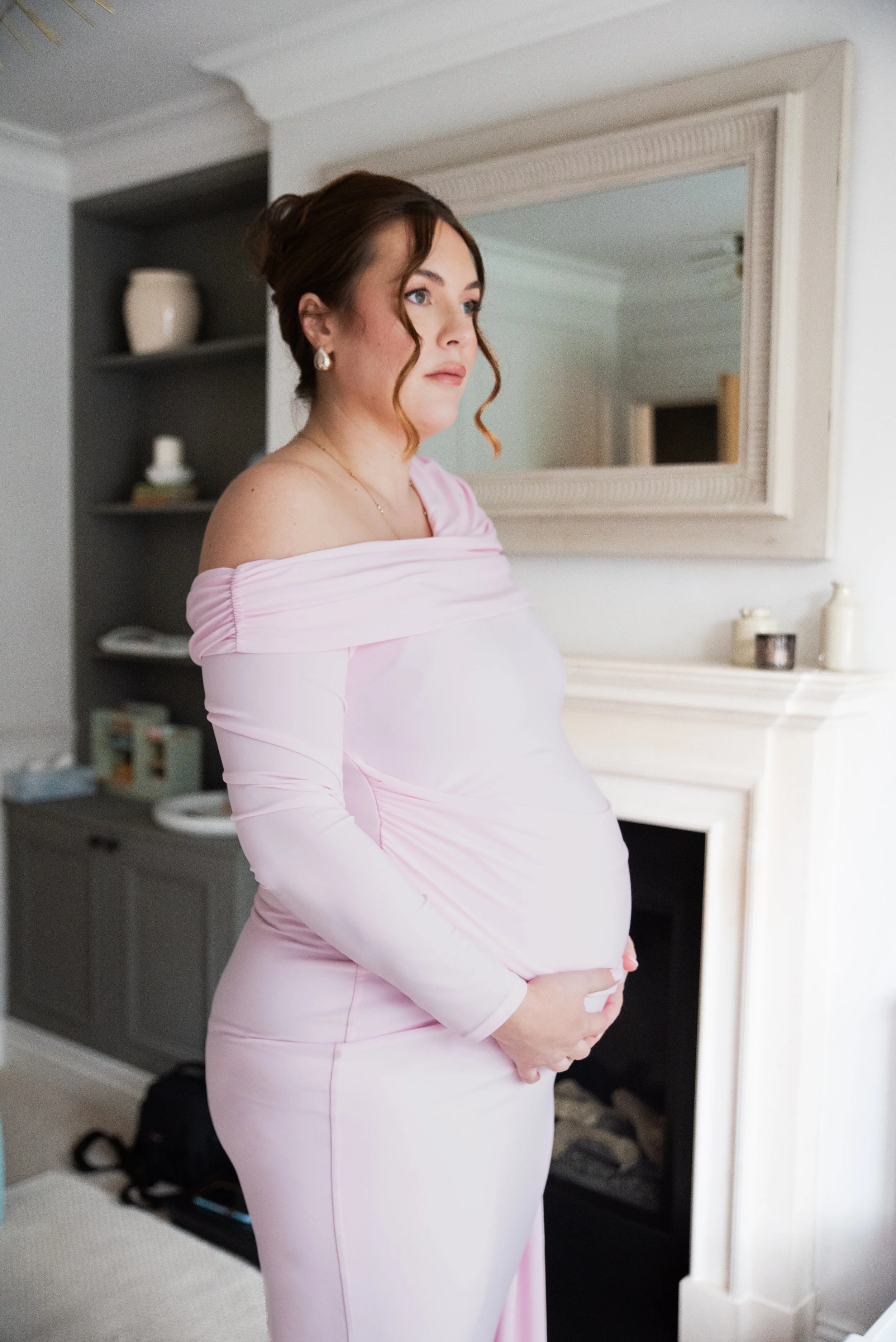 Pregnant woman in a pink off-shoulder dress standing like she's waiting, holding her belly near a white fireplace in a cozy room with a large mirror.