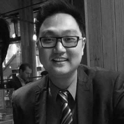 Smiling man wearing glasses and a suit indoors, with blurred people in the background.