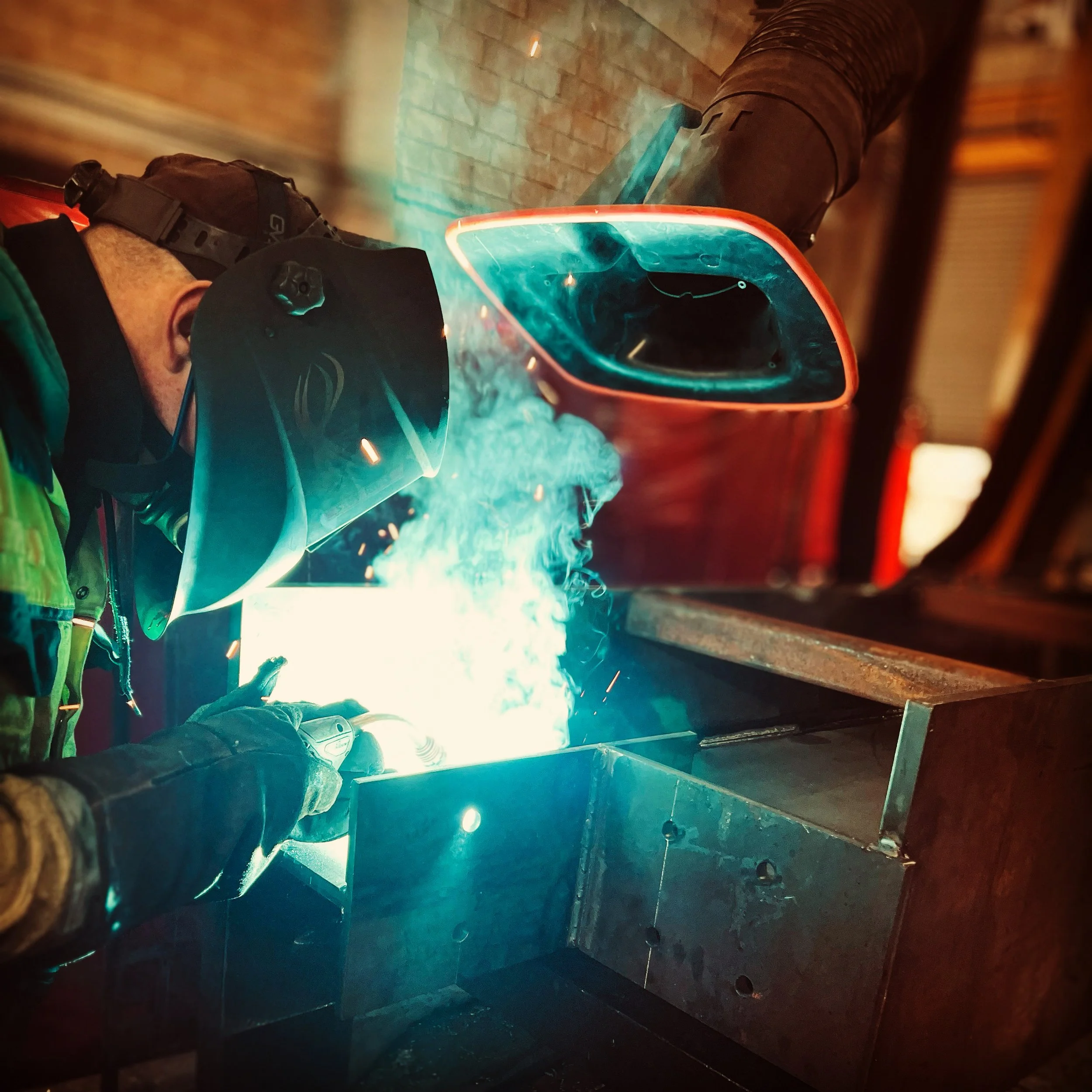 A worker welding metal, wearing a helmet and protective gear, with sparks and blue light from the welding process.
