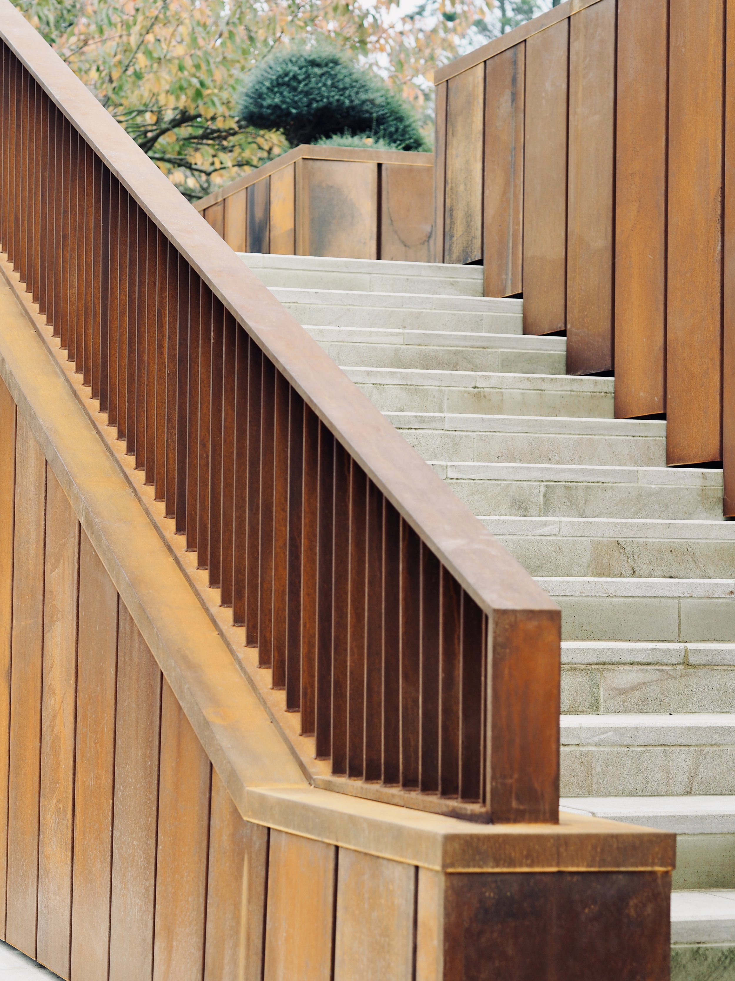 Concrete stairs with corten railing on a building's exterior, with trees in the background.