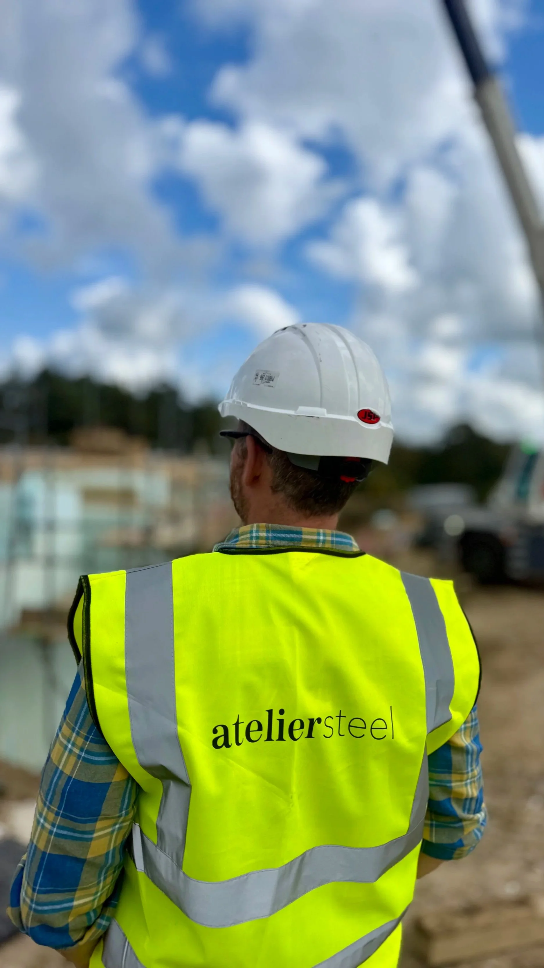 A construction worker wearing a white hard hat and bright yellow safety vest with the words 'ateliersteel' on the back, standing on a construction site with a cloudy sky overhead.
