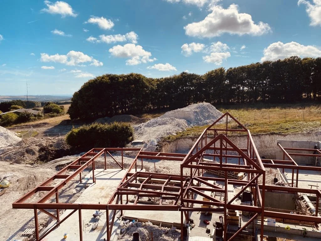 Construction site with steel framing, piles of dirt, and trees in the background under a partly cloudy sky.