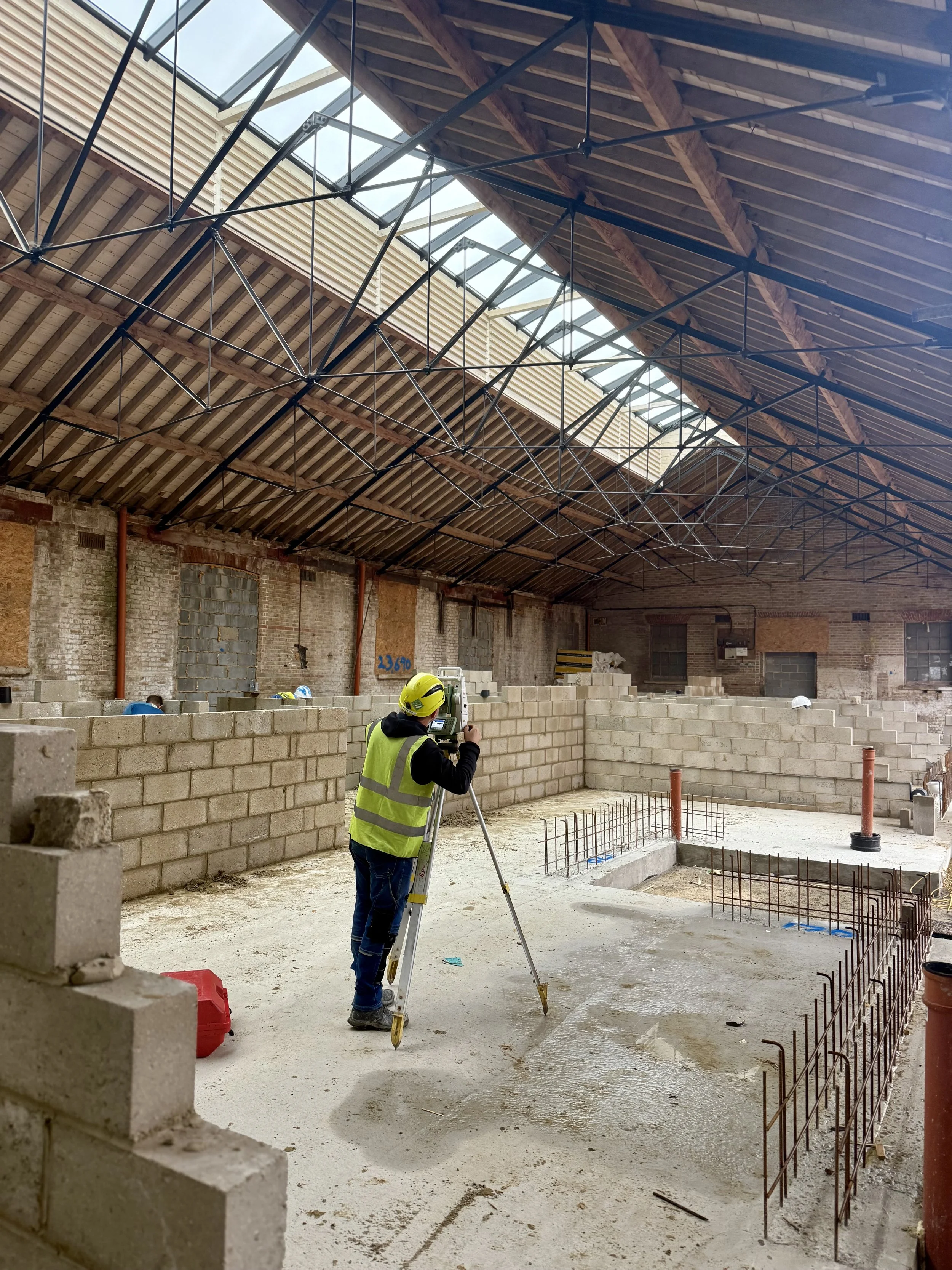Construction worker in a yellow safety vest and helmet using a leveling instrument inside a large building under construction with brick walls, rebar, and a roof with skylights.