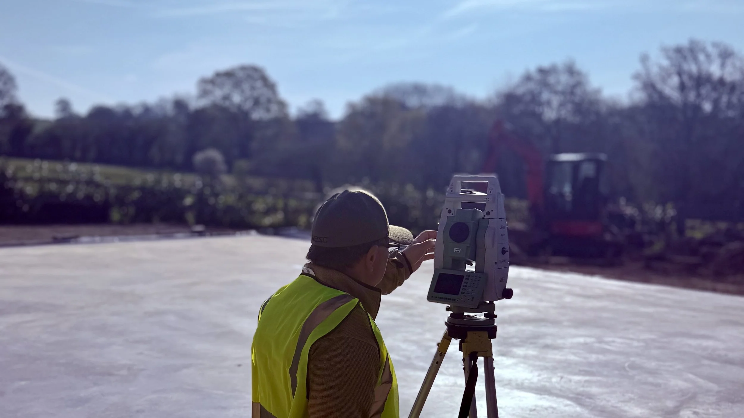 A construction worker wearing a yellow safety vest and a cap is using surveying equipment on a tripod at a construction site, with trees and construction machinery in the background.