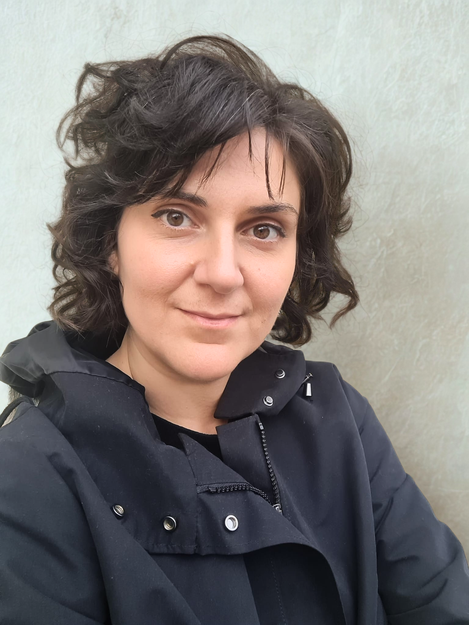 A woman with short, curly dark hair and brown eyes, wearing a black jacket, smiling while taking a selfie against a plain light-colored wall.