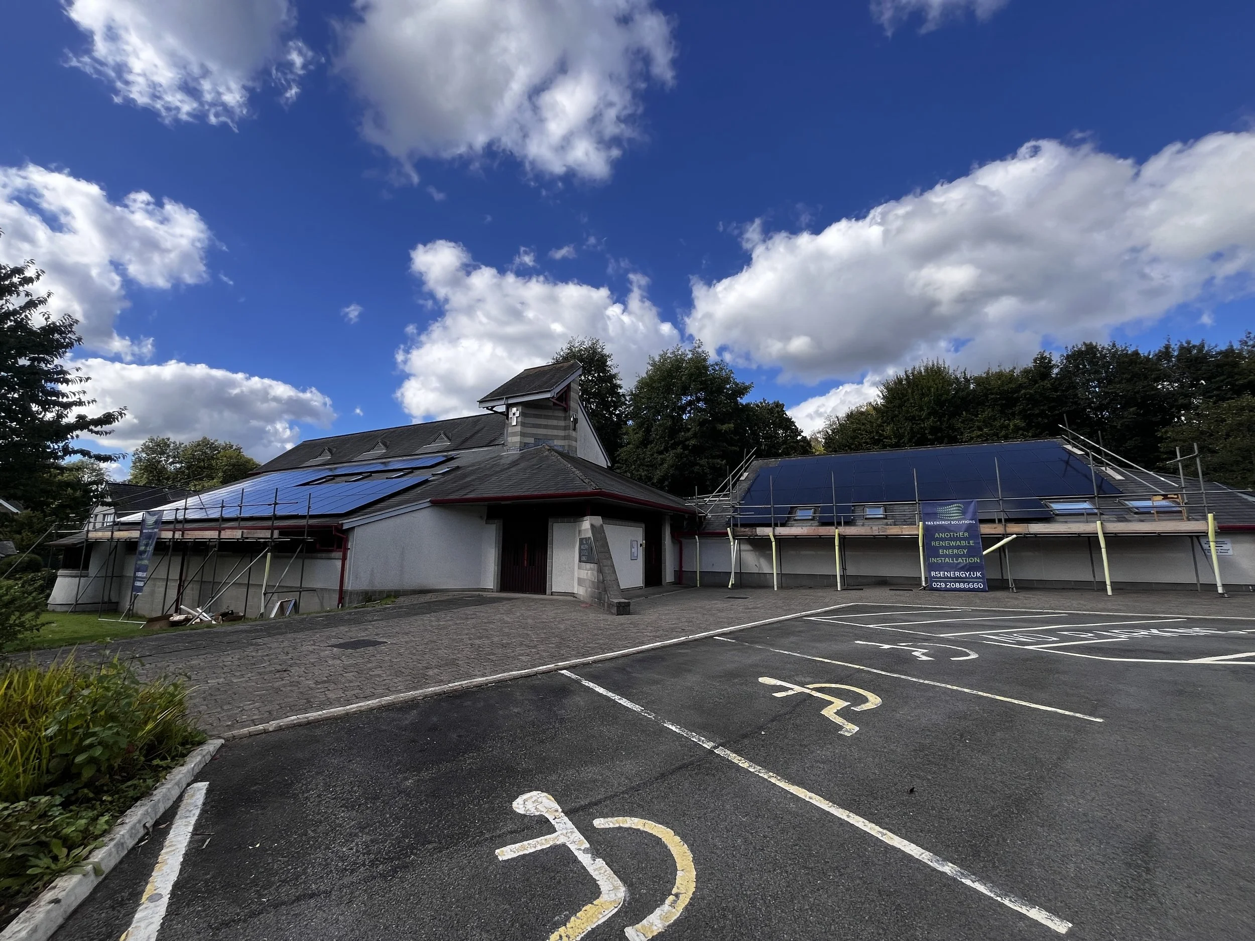 Building with solar panels on roof, parking lot with designated handicapped parking spaces, trees, and a partly cloudy sky.