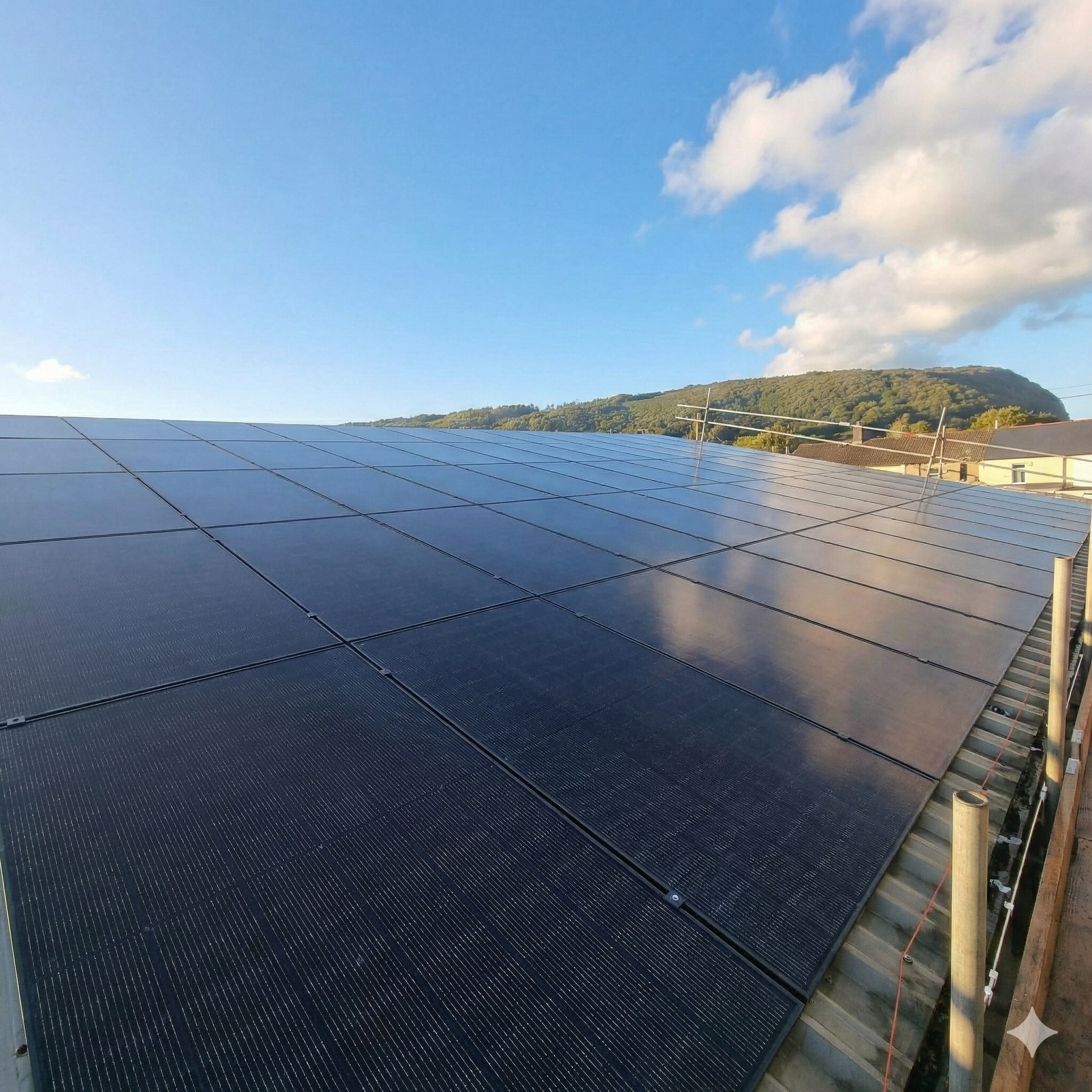 A rooftop with numerous solar panels installed, with a background of a hilly landscape and blue sky with some clouds.