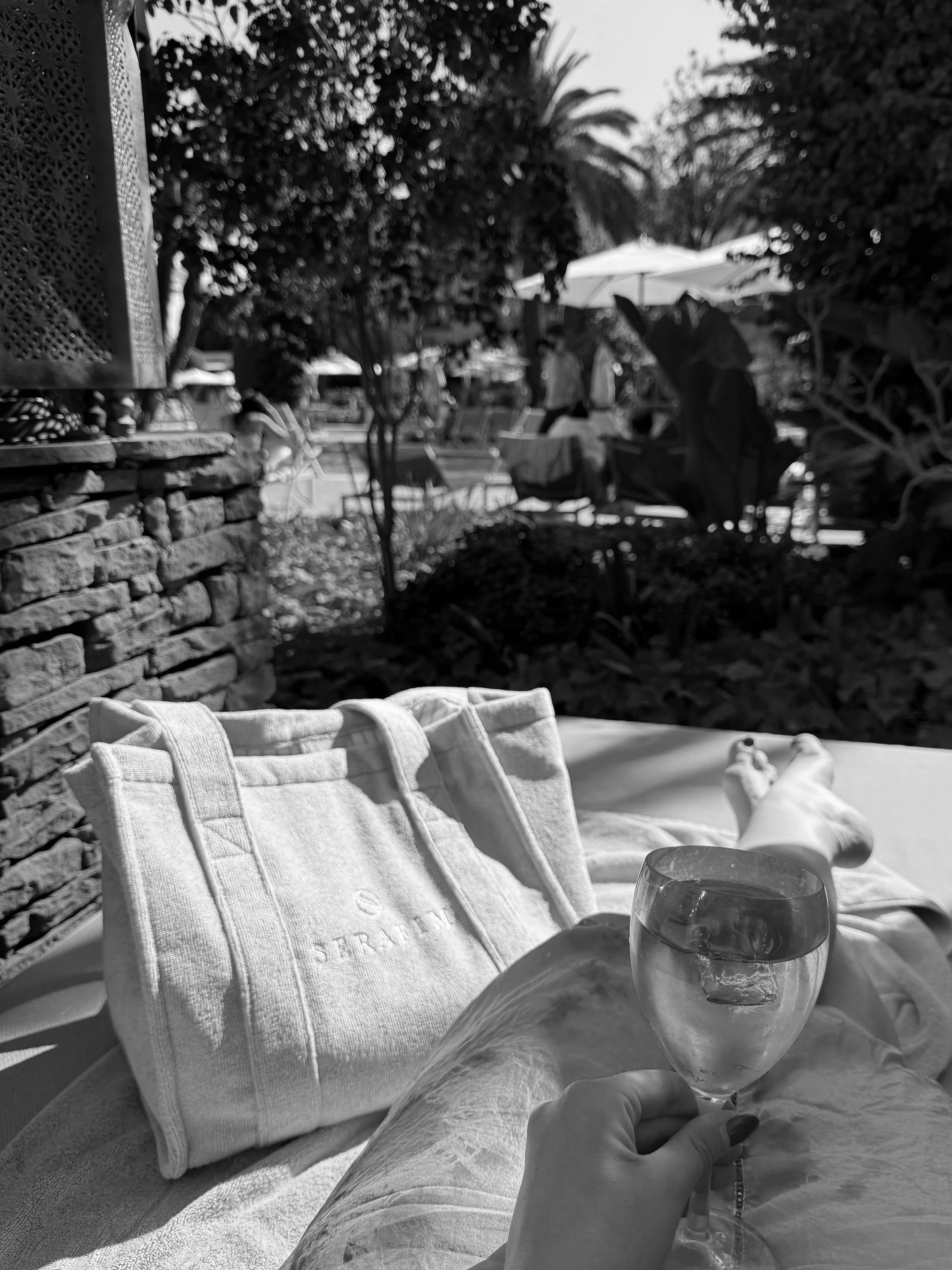 A person lounging outdoors on a sunny day, holding a glass of water or clear beverage. Their legs are stretched out on a lounge chair. Nearby are a tote bag labeled 'STRADE' and a textured towel. The background features trees, umbrellas, and poolside