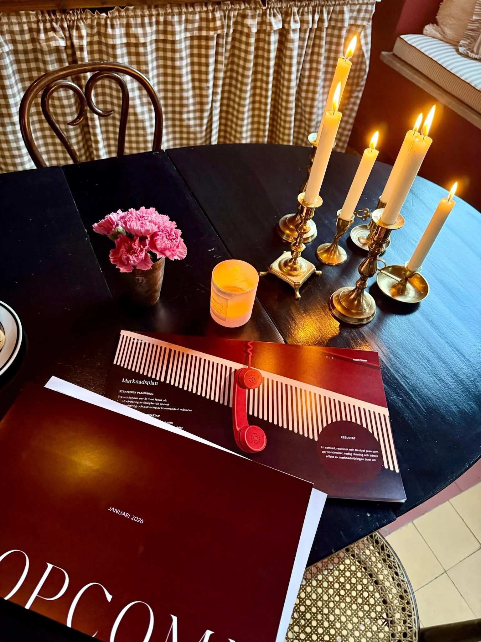 A dining table with pink flowers in a pot, lit candles in brass holders, a red paper with a red vintage telephone, and a planner, with a checkered curtain in the background.
