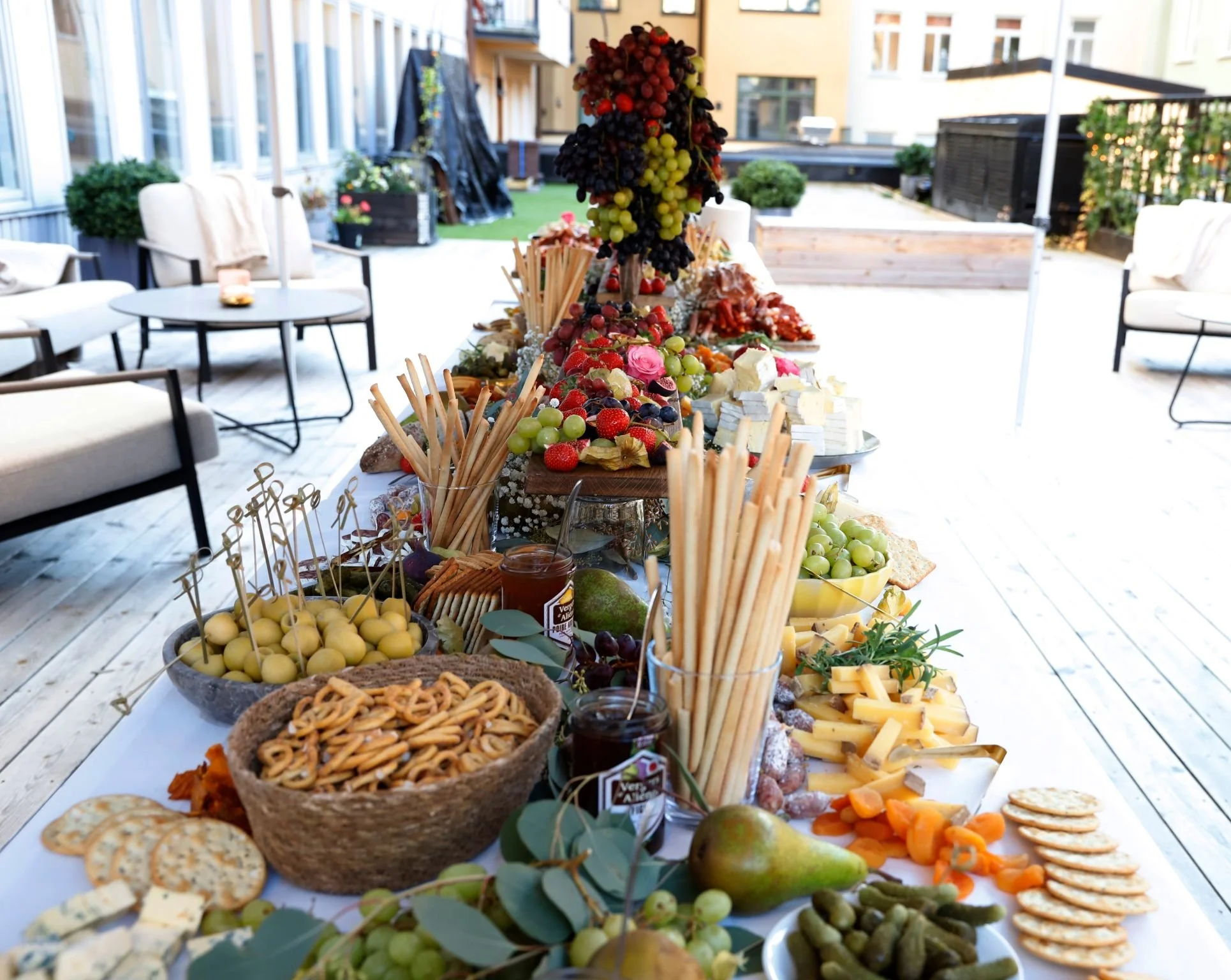 A long outdoor table set with various cheeses, fruits, crackers, and snacks at a gathering, with patio furniture and a cityscape in the background.