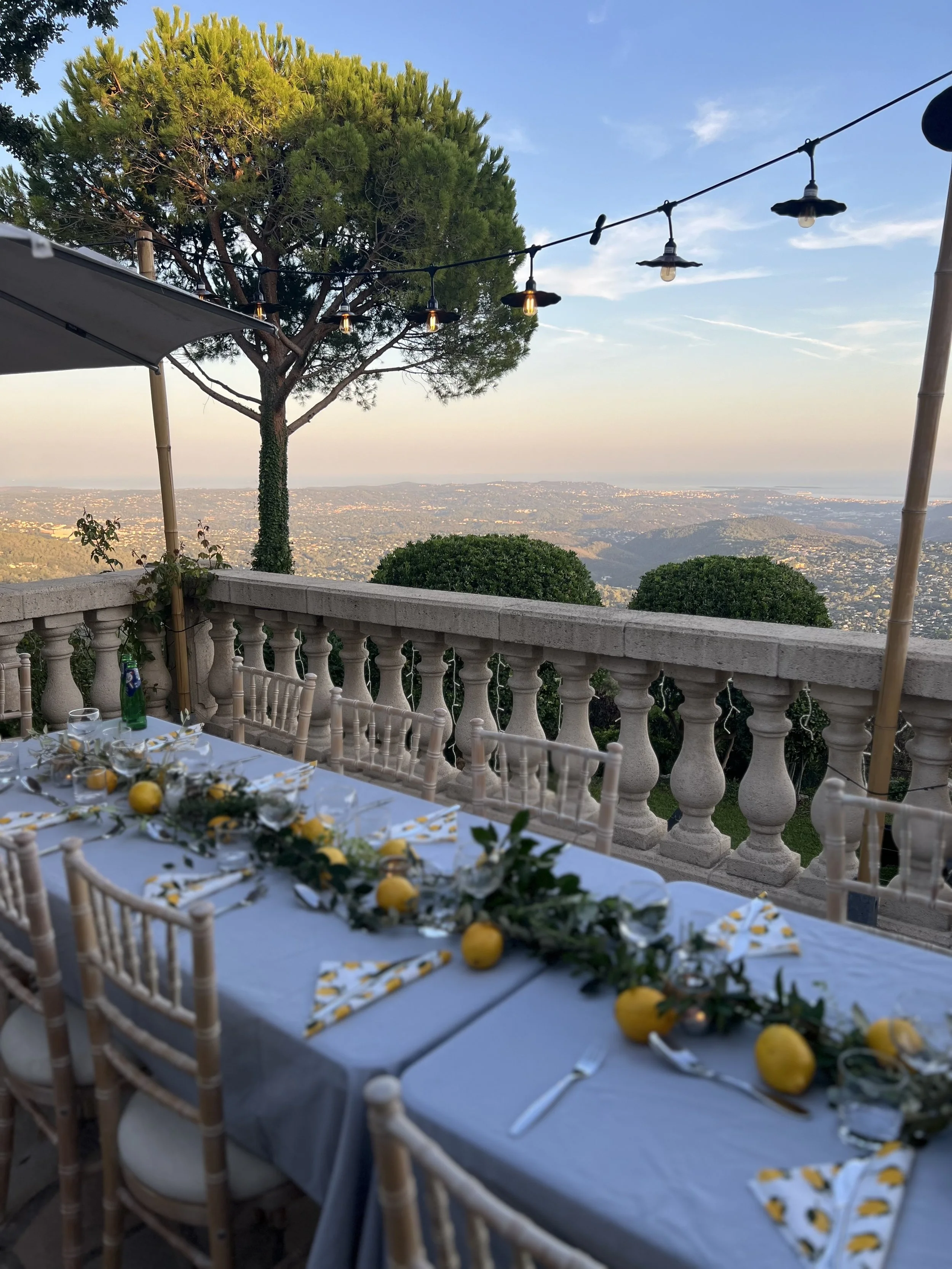 Outdoor dining setup with a long table decorated with lemons and greenery, overlooking a scenic view of hills and a cityscape at sunset, with string lights hanging overhead.