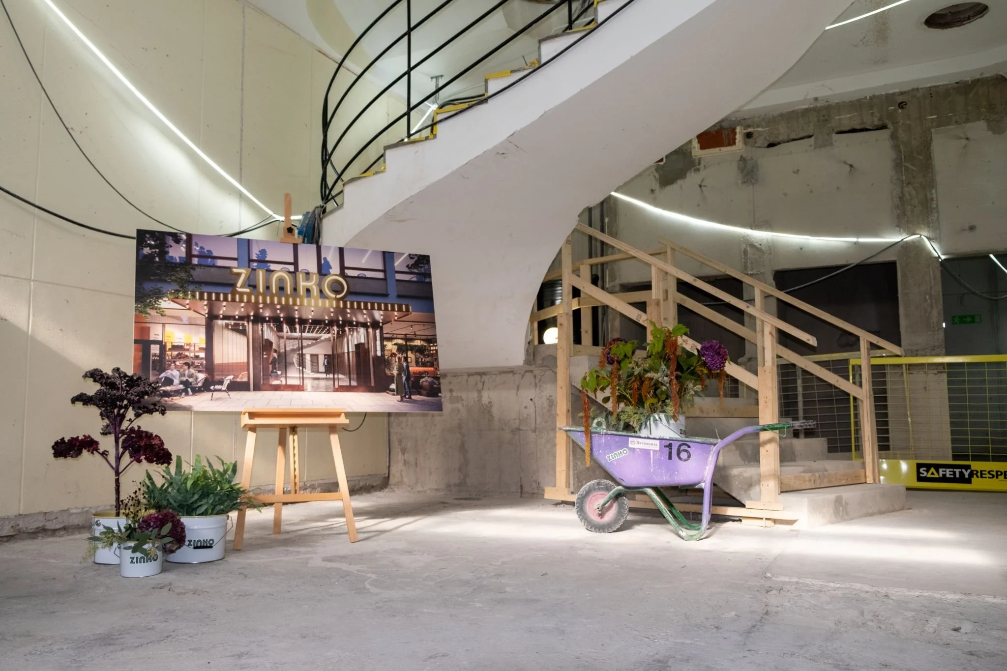 Unfinished indoor space with construction materials, potted plants, a cart with flowers, a sketch of a building on an easel, staircase with railings, and a safety sign.