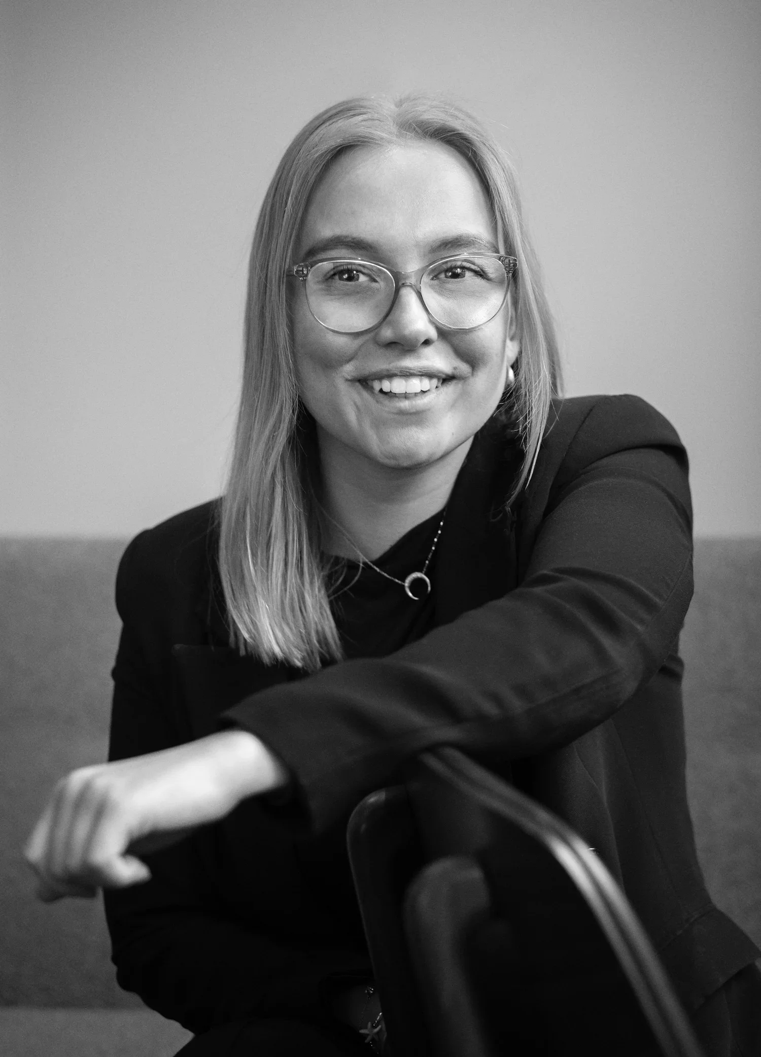 Black and white photo of a smiling young woman with glasses and long hair, sitting on a chair, wearing a dark blazer and jewelry, in a simple indoor setting.