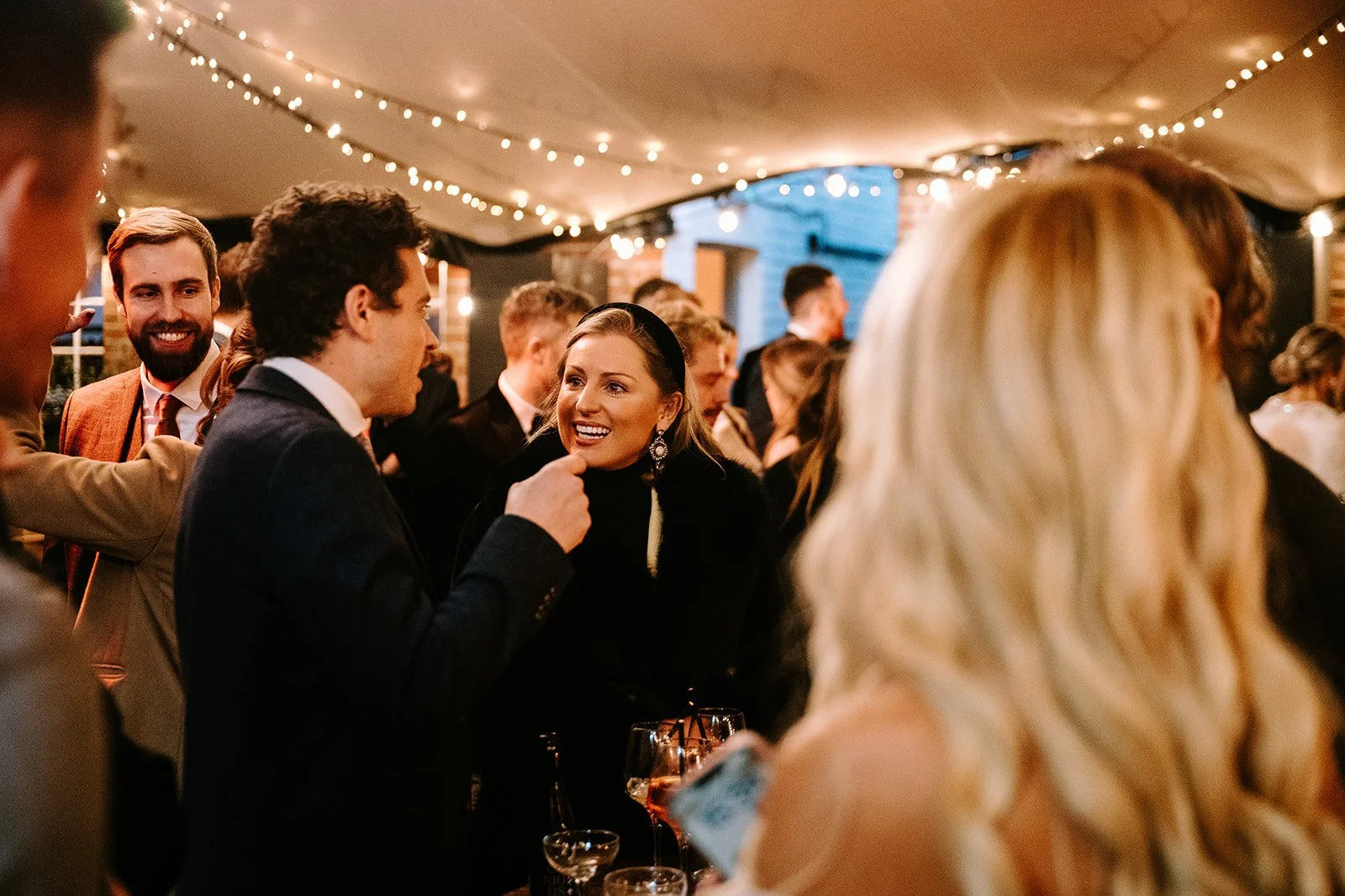 People socializing at a gathering under string lights, smiling and talking in a warmly lit indoor setting.