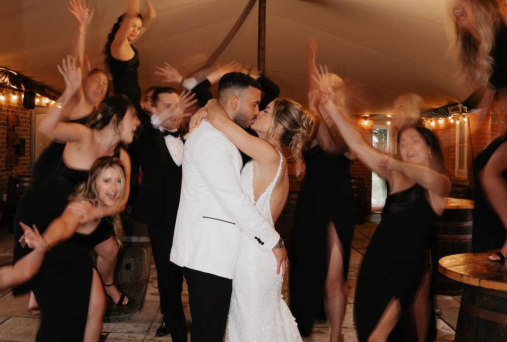 Bride and groom kissing with bridesmaids celebrating around them in a dimly lit venue.