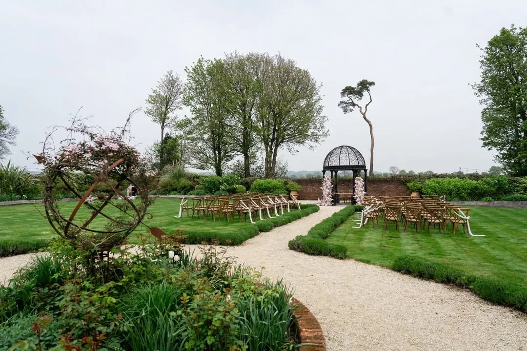 Outdoor garden wedding setup with rows of chairs on either side of a grassy aisle leading to a decorative gazebo with floral arrangements.