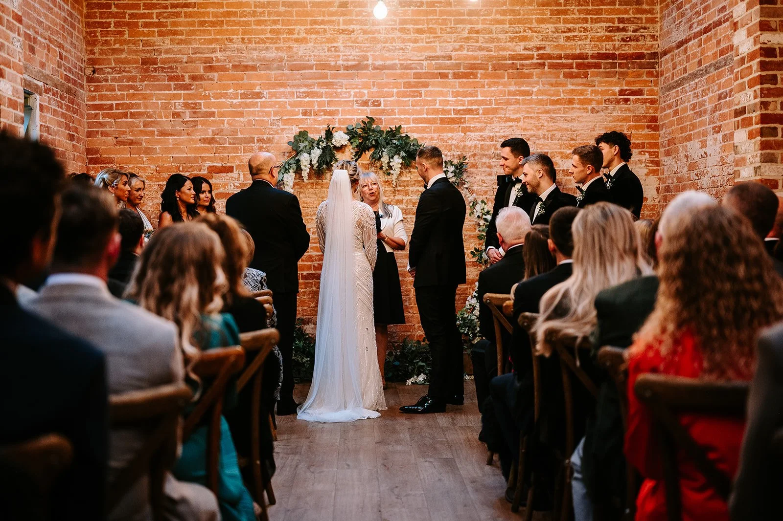 A wedding ceremony with a bride and groom exchanging vows in front of a officiant, standing against a brick wall decorated with green foliage and white flowers, while guests watch.