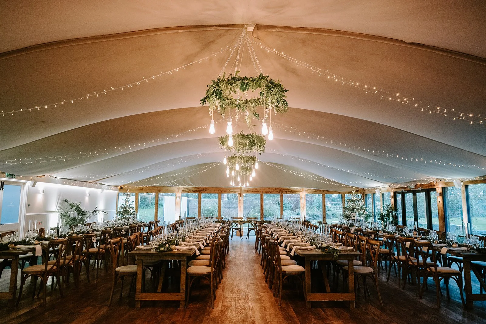 Elegant event space with long wooden tables and chairs, decorated with candles, flowers, and greenery, illuminated by hanging light bulbs and string lights, with large windows showing an outdoor view.