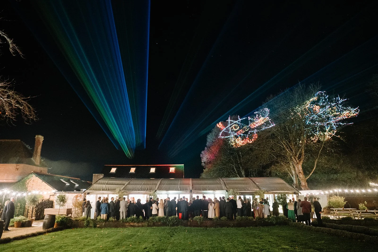 A nighttime outdoor event with a crowd gathered in front of a building decorated with string lights. There are colorful laser beams projecting into the sky and illuminated festive drawings on a tree.