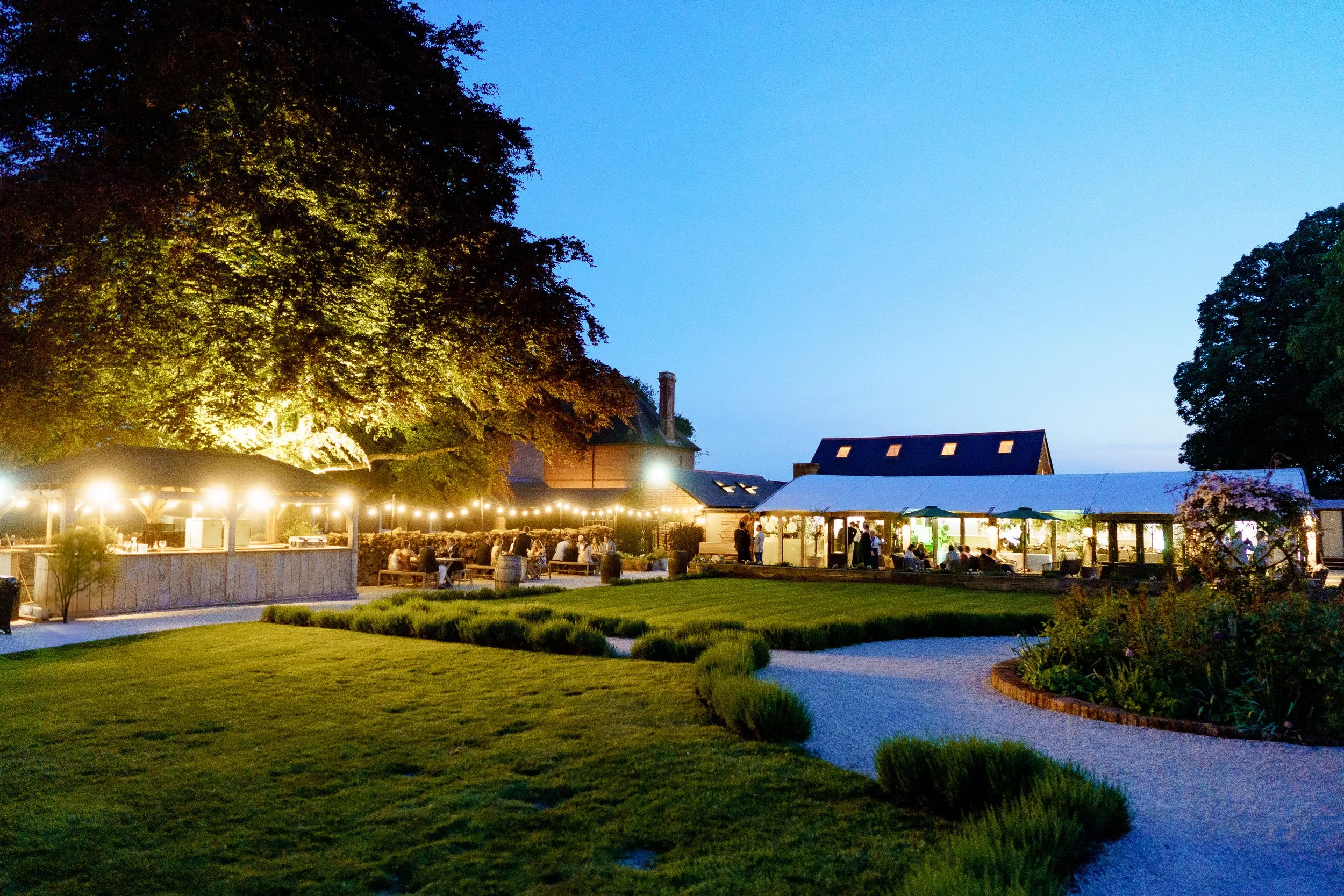 Outdoor evening restaurant with illuminated string lights, people dining under a tent, large trees, manicured lawn, and garden.