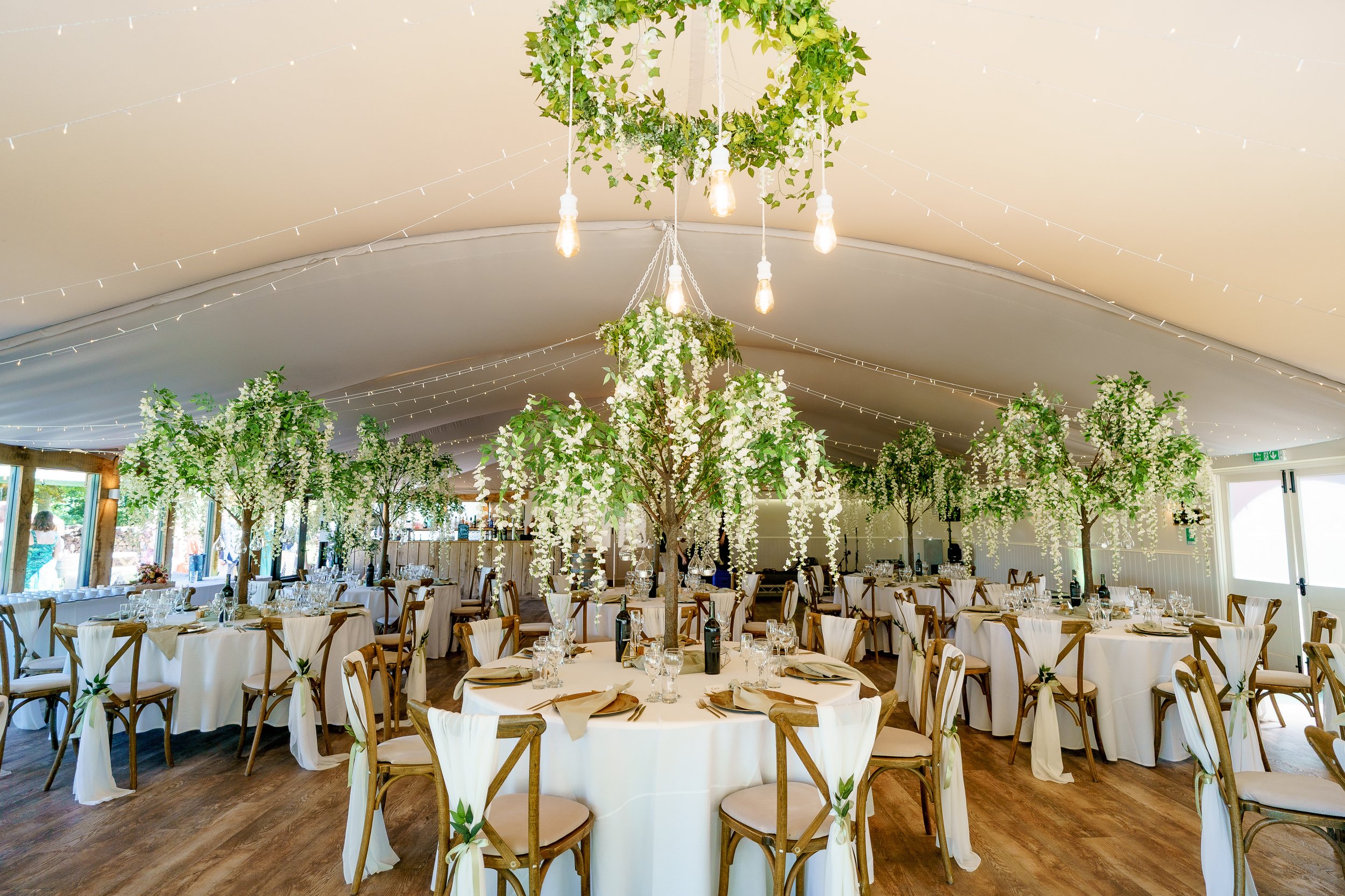 Elegant event tent decorated with hanging and potted greenery, featuring round tables with white tablecloths, chairs with white tied sashes, and centerpieces with white flowers and wine bottles.