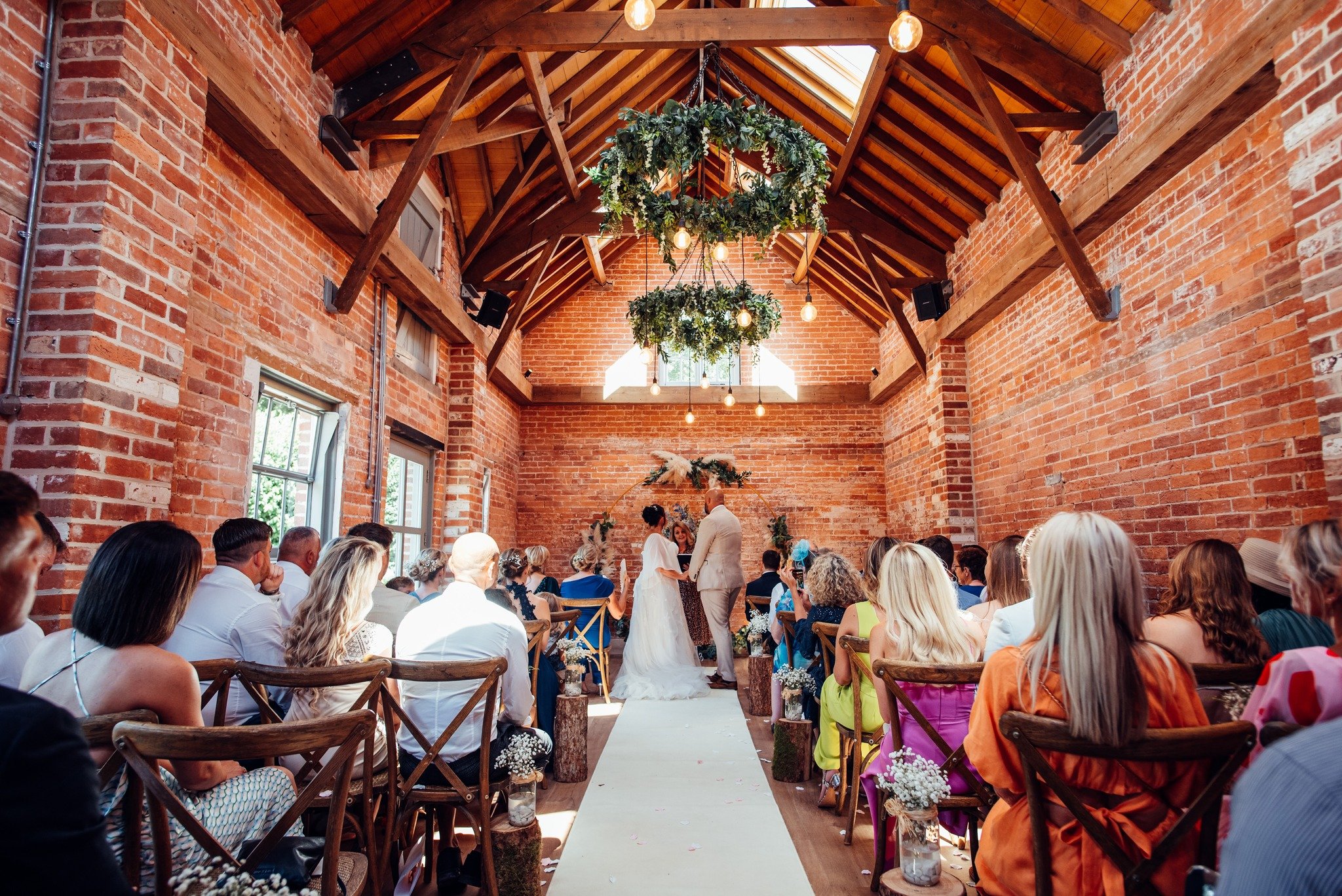 Indoor wedding ceremony in a rustic venue with brick walls, wooden beams, floral decorations, and guests seated in wooden chairs.