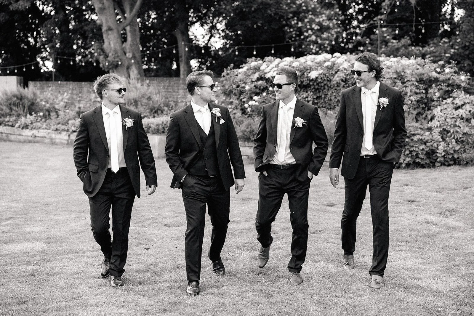 A group of five men in suits and sunglasses walking on a grassy lawn, smiling and talking to each other, with trees and bushes in the background.