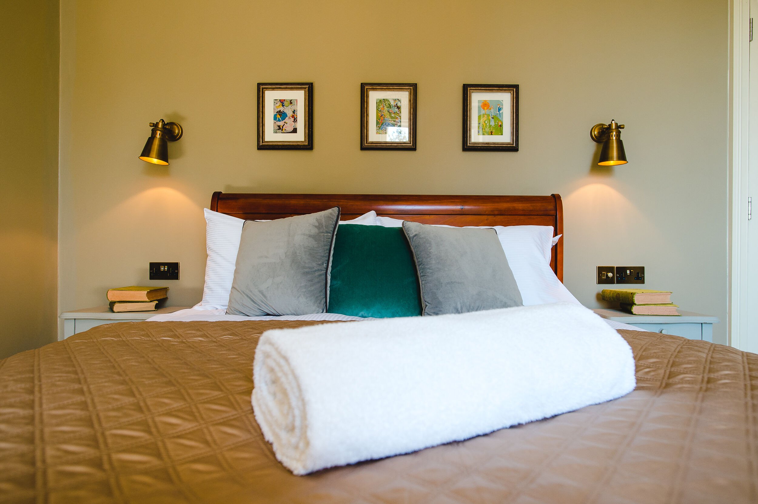 Cozy bedroom with a bed featuring gray and teal pillows, a brown quilt, and a rolled white towel. Wall-mounted lamps and framed artwork adorn the wall above the wooden headboard.