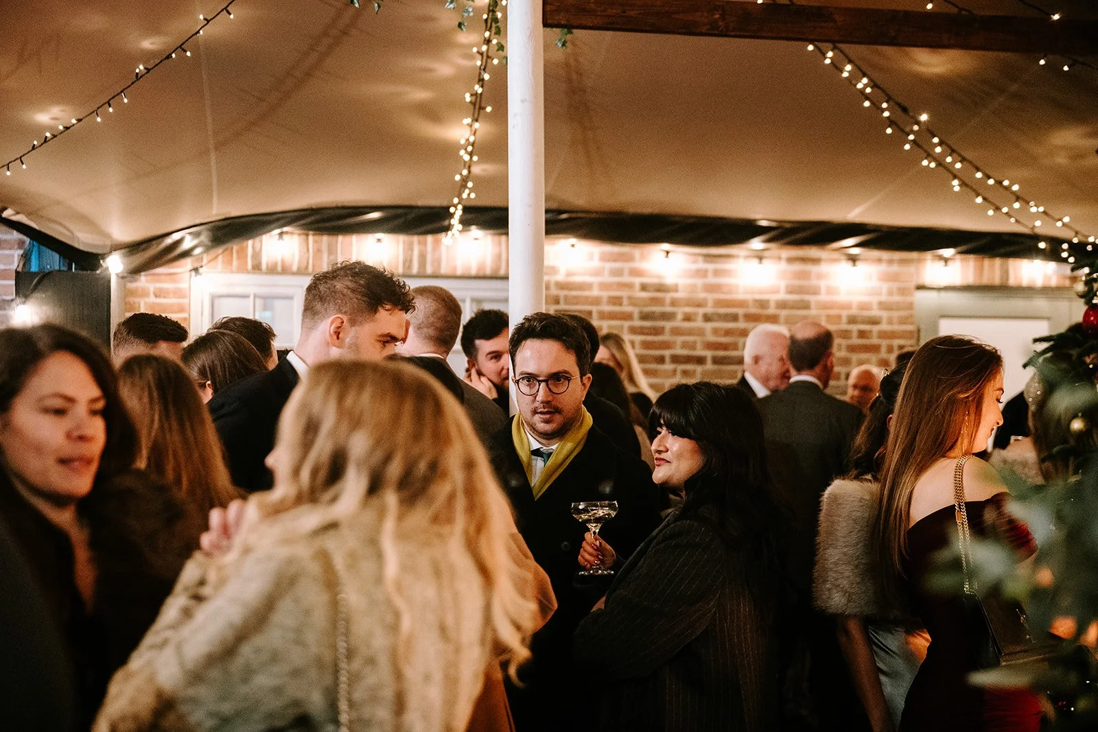 People socializing at a festive house party under string lights with a brick wall background.
